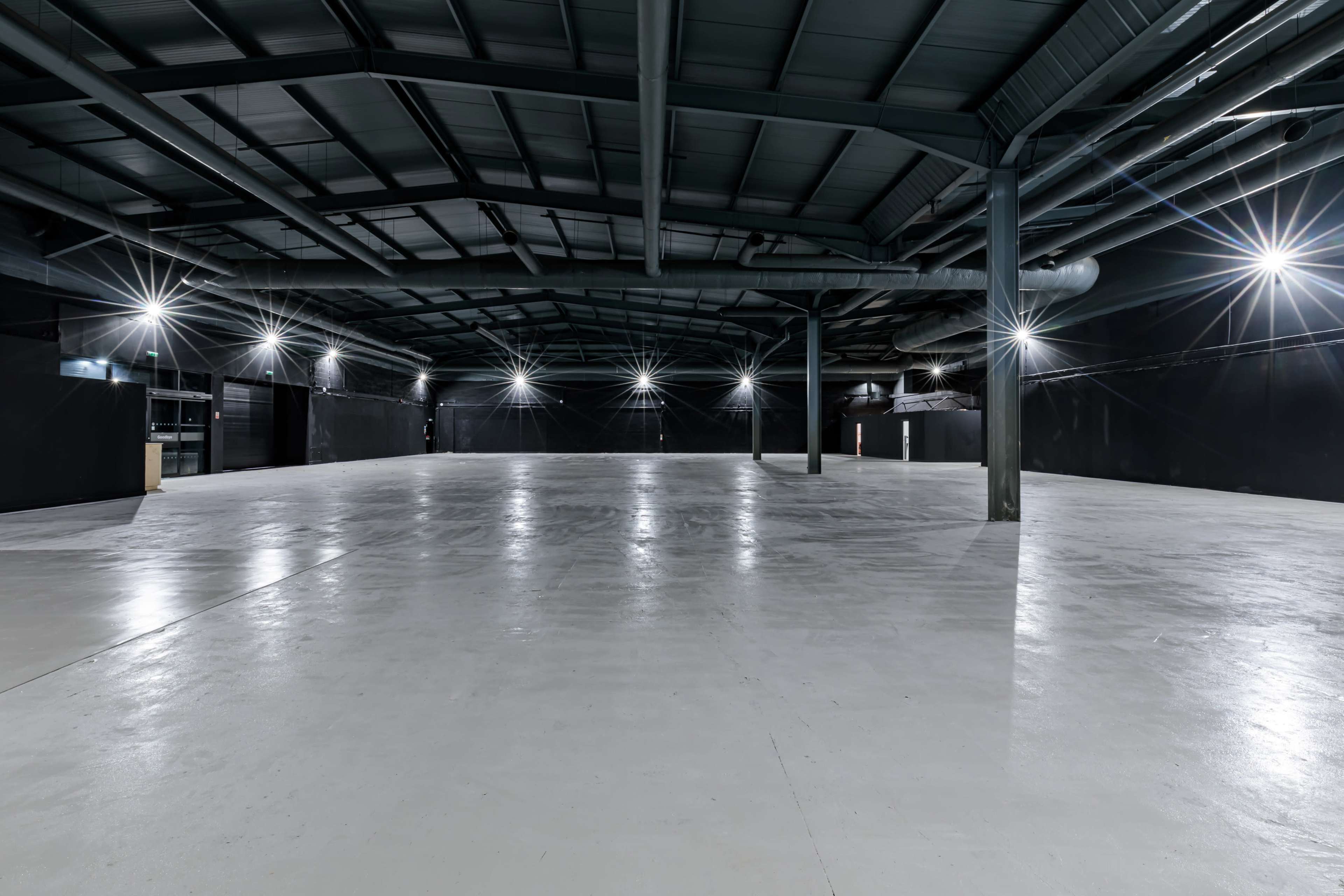 The image shows a large, empty industrial space with a high ceiling and bright overhead lights illuminating the concrete floor.