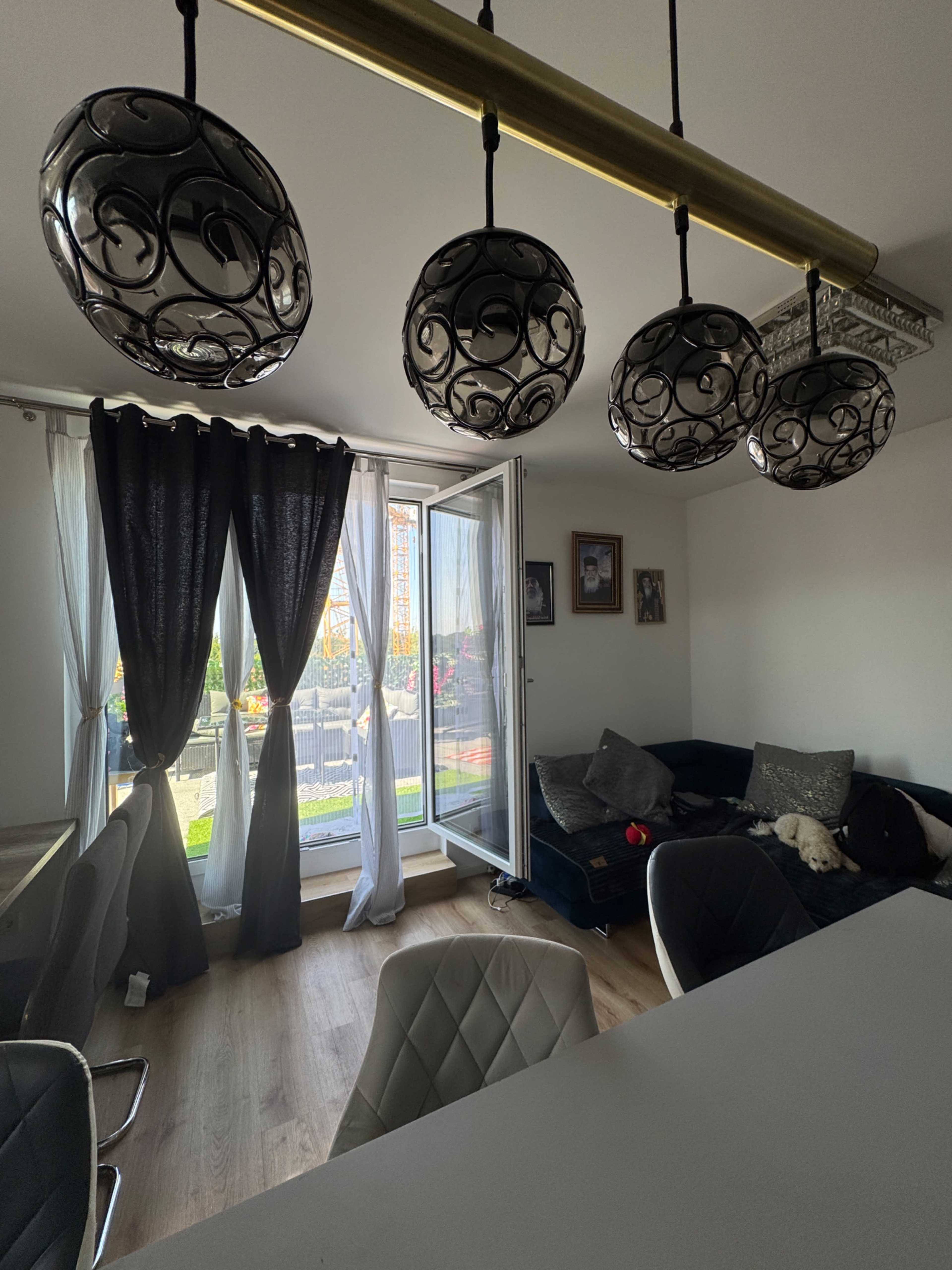 A modern living room features a dark-colored couch, a dining area with light-colored chairs, decorative hanging lights, and a balcony with a view.