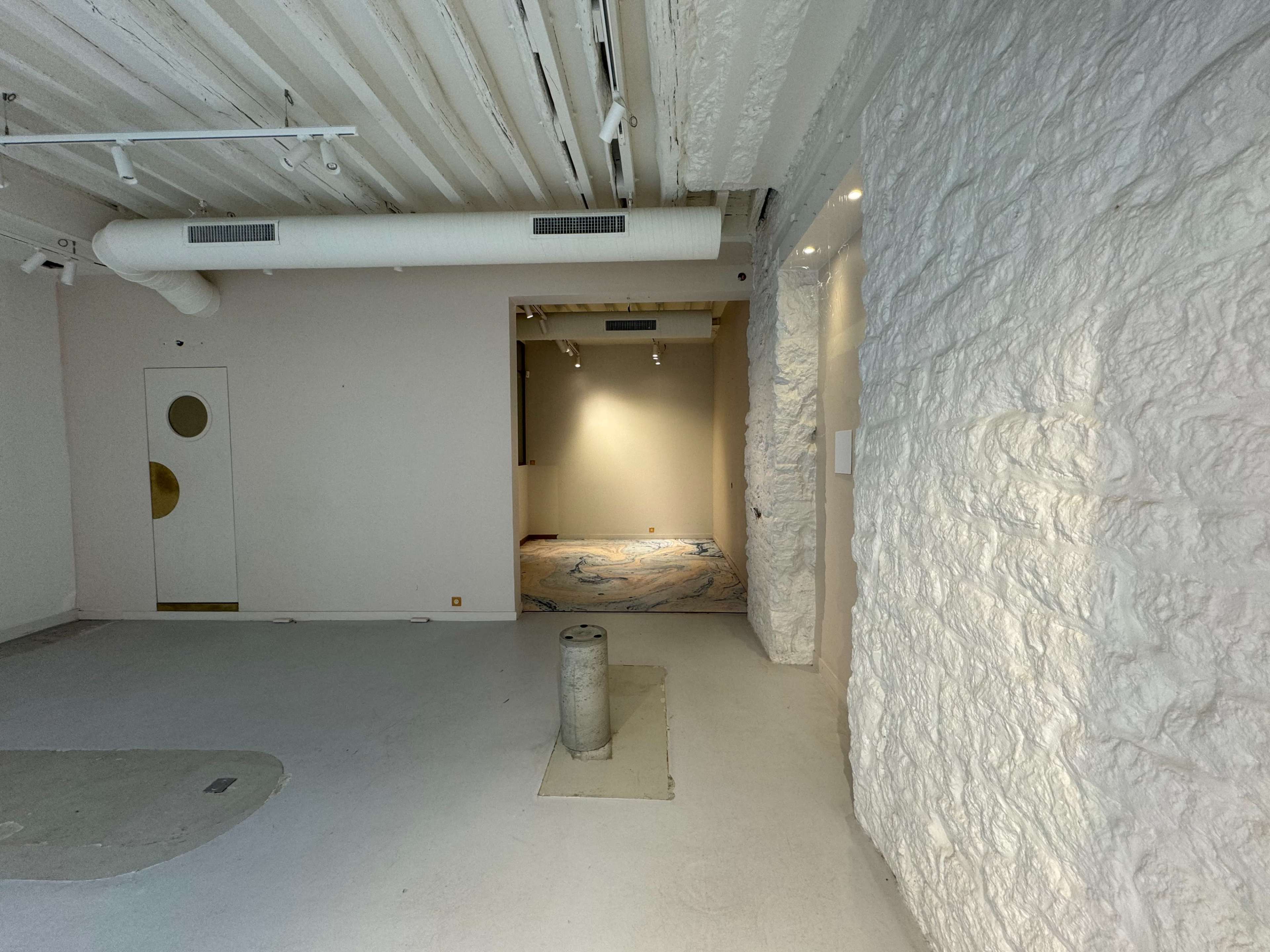 An empty room with a textured white wall, a circular doorway, and an adjoining room illuminated by overhead lights.