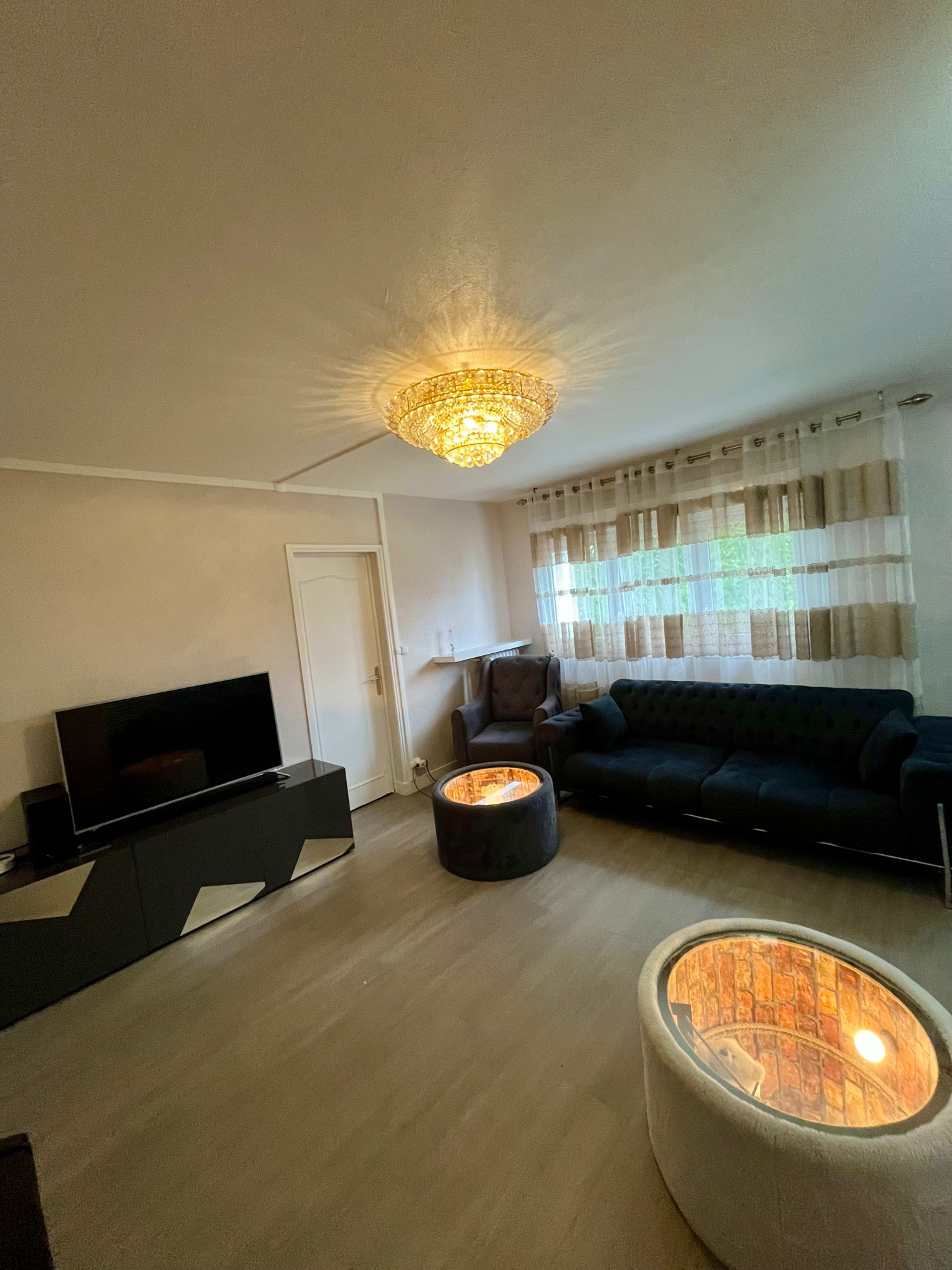 The living room features a black sofa, a television stand, and two circular coffee tables with illuminated interiors under a chandelier and large windows covered with sheer curtains.