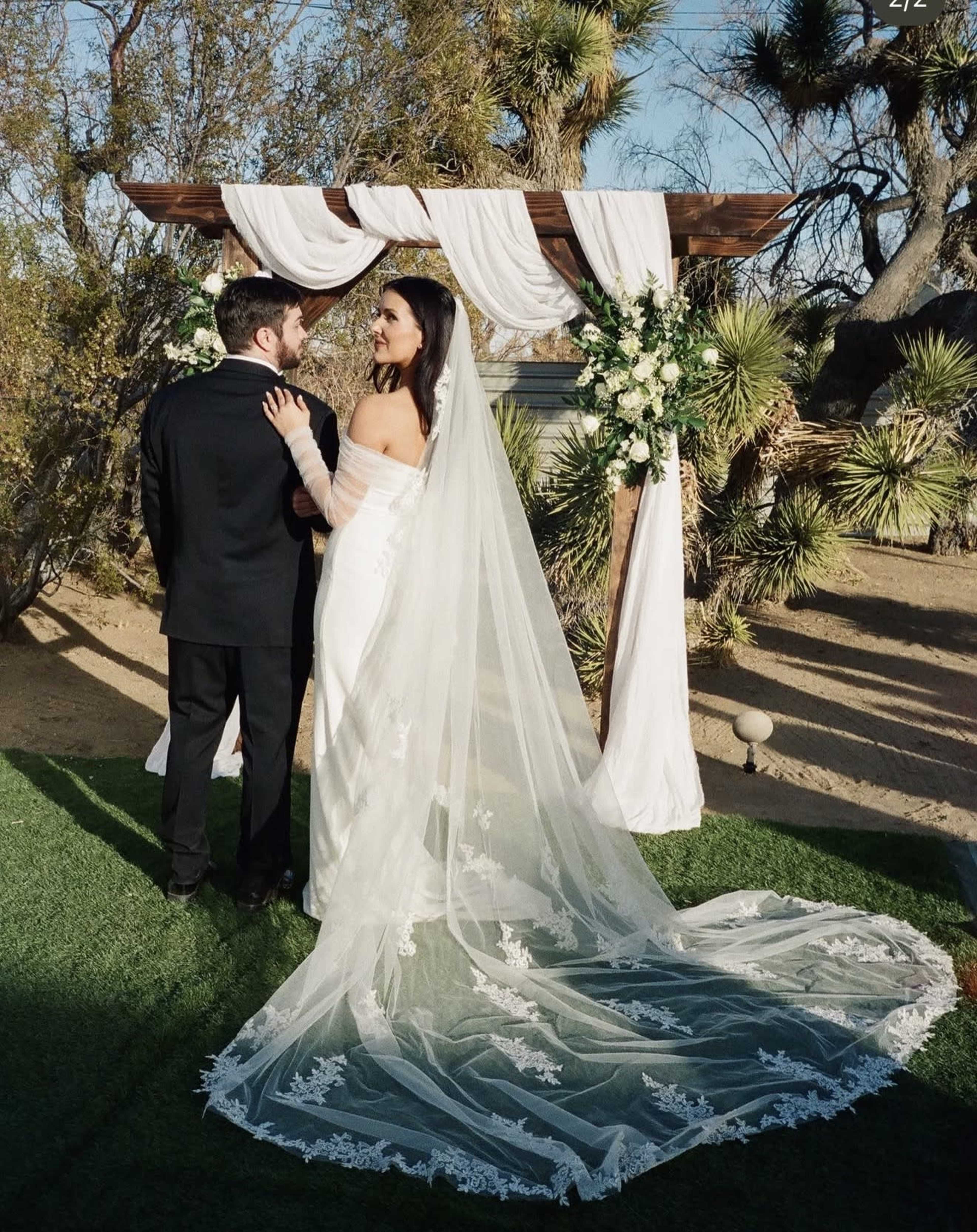 A couple stands beneath a decorated wedding arch, with the bride in a white gown and veil and the groom in a black suit, surrounded by desert vegetation.