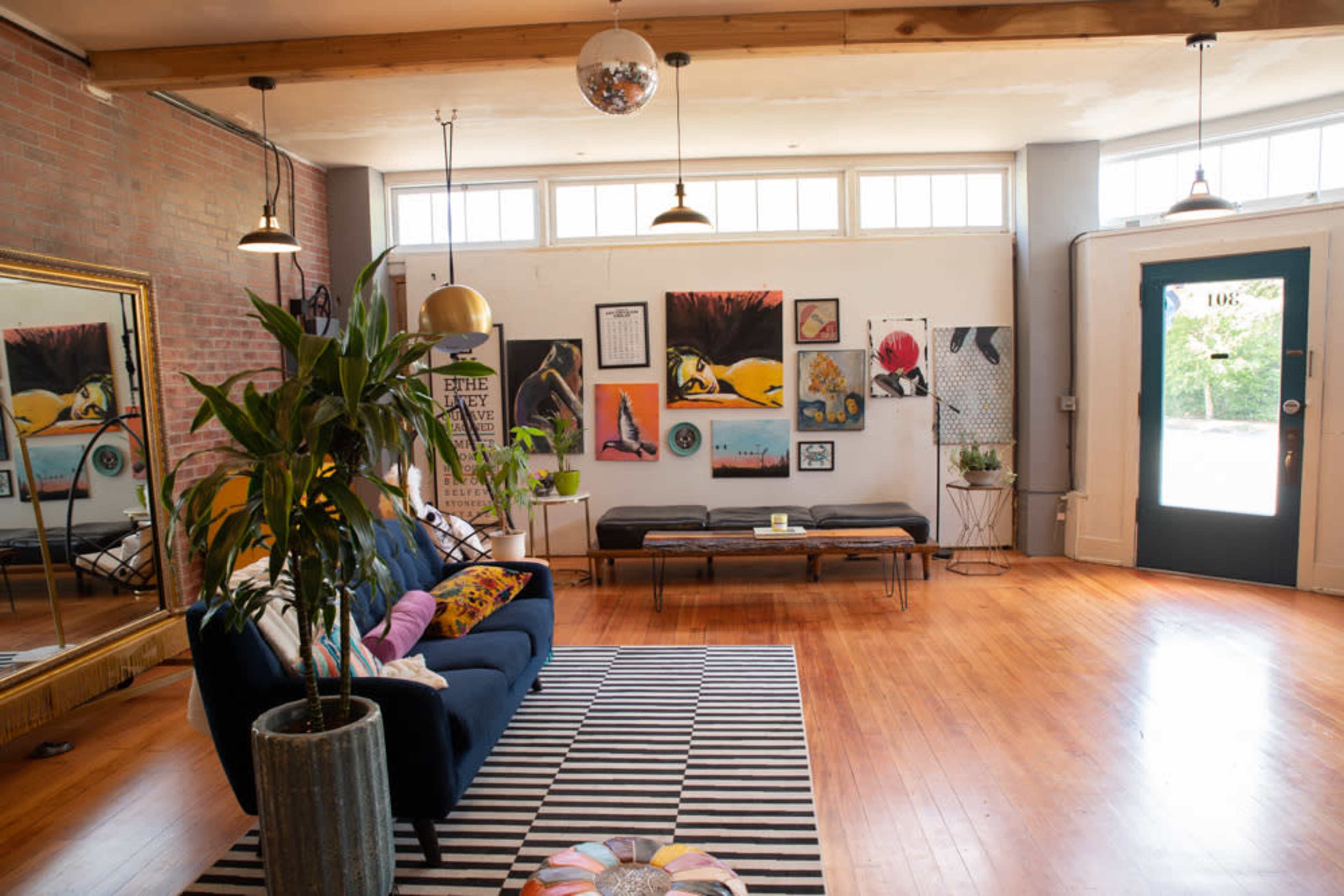 The image shows a bright living area featuring a blue sofa, a striped rug, and various art pieces on the walls, illuminated by natural light from large windows.