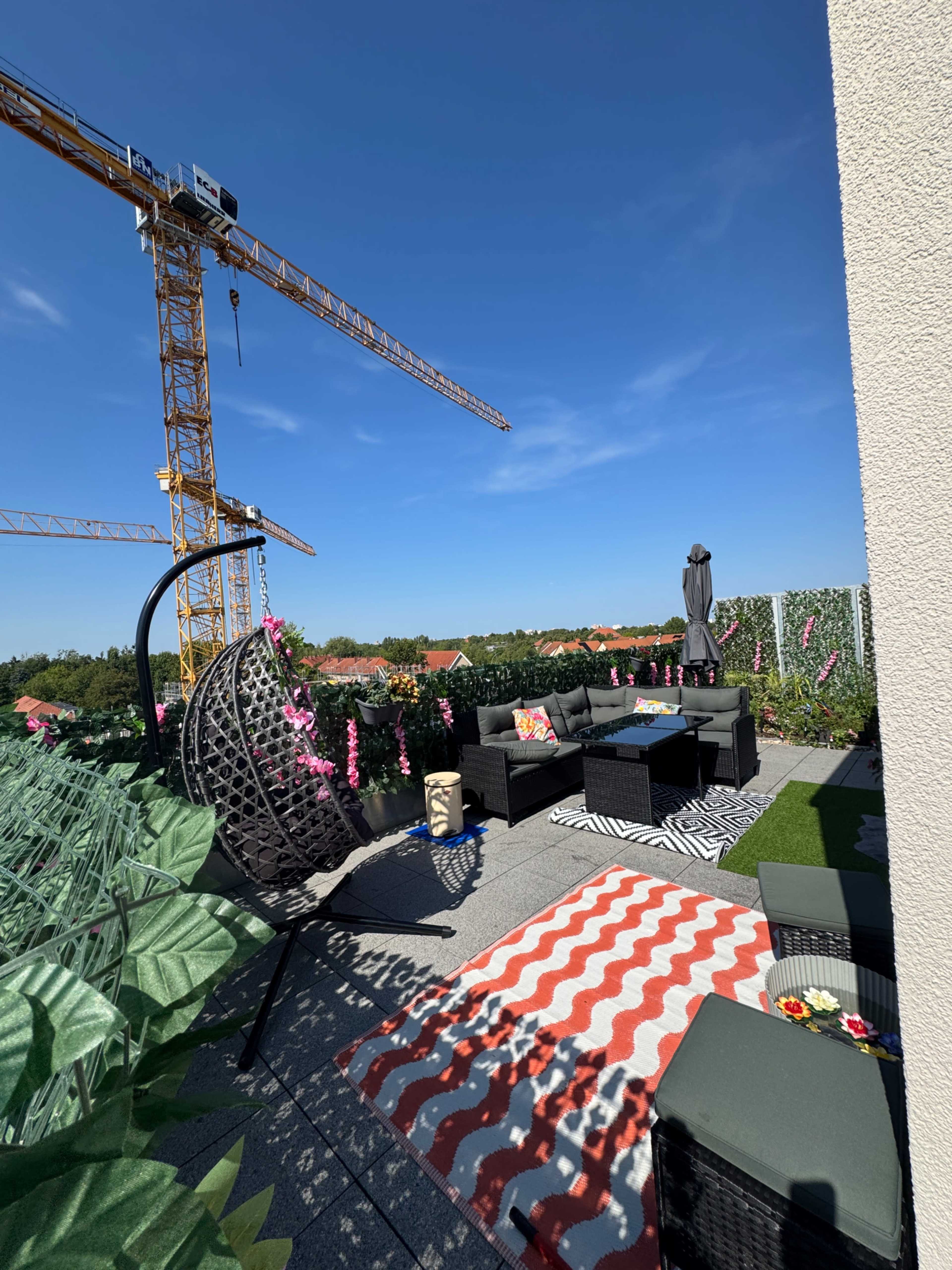 The image shows a rooftop terrace featuring black outdoor furniture, decorative rugs, potted plants, and a construction crane in the background.