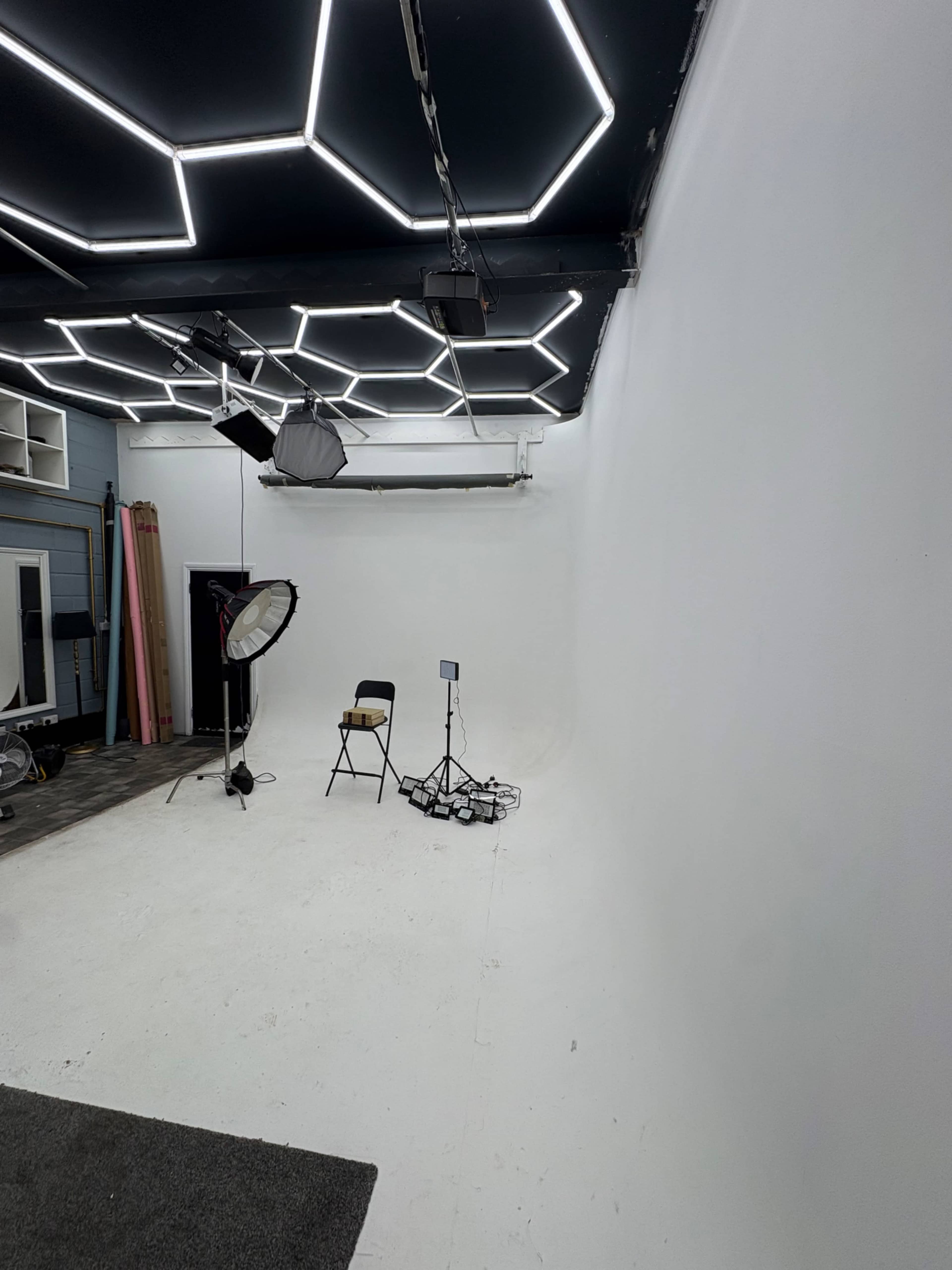 The image shows an empty photo studio with a white backdrop, a single chair, and various lighting equipment.