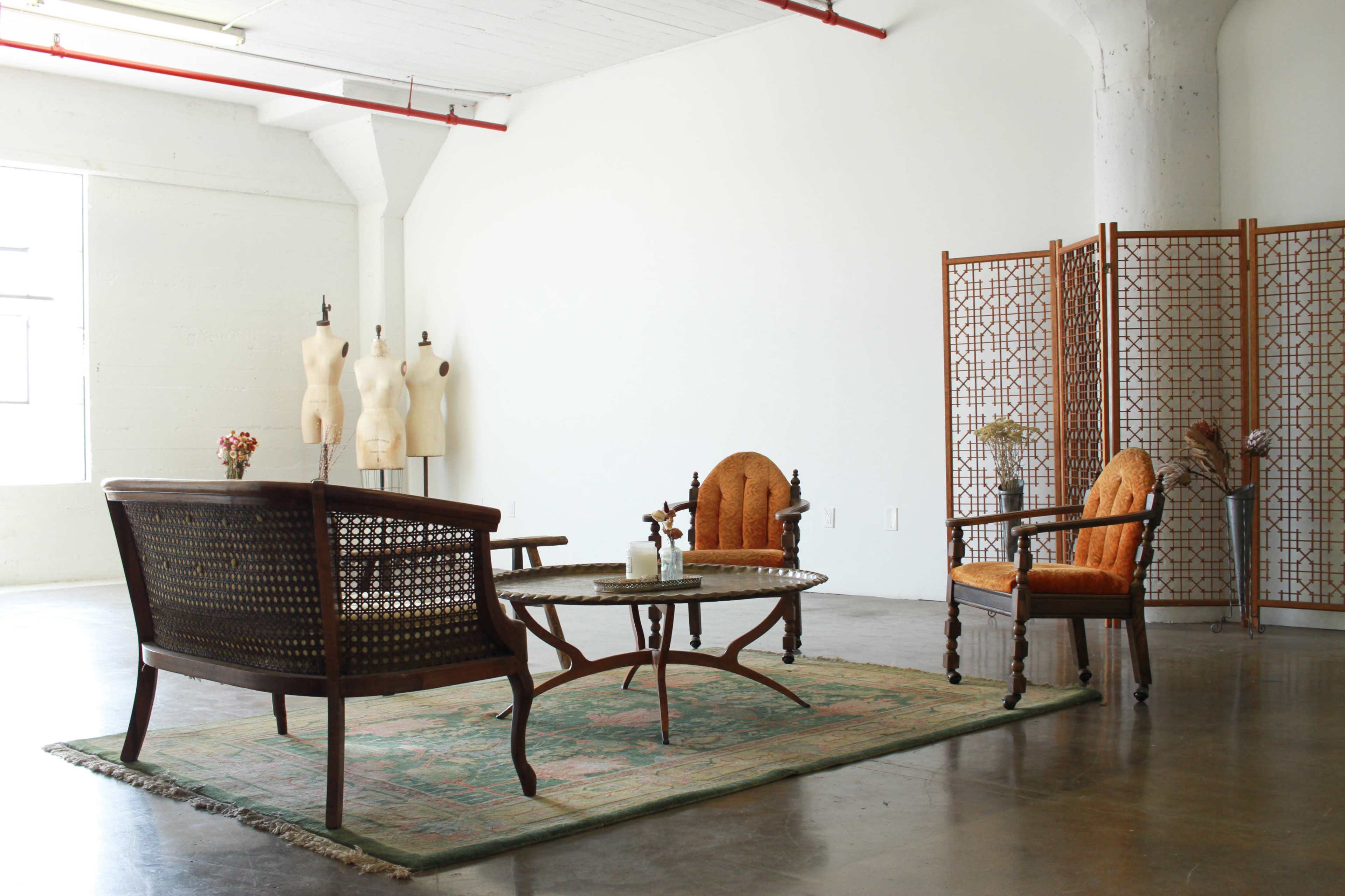 A minimalist room with a wooden seating arrangement, a round table, and decorative screens, complemented by a vintage area rug and dressmaker forms in the background.