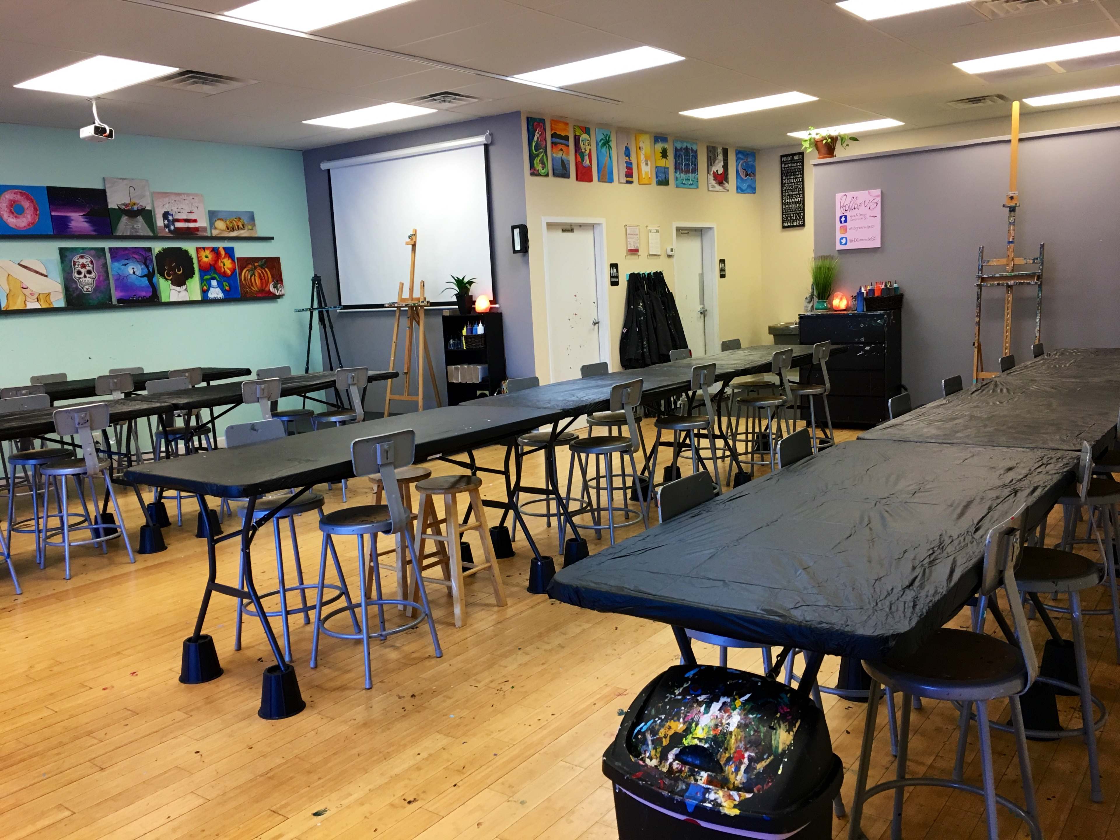 An art studio with several long tables covered in black tablecloths, surrounded by tall stools, and walls adorned with colorful artwork and supplies.