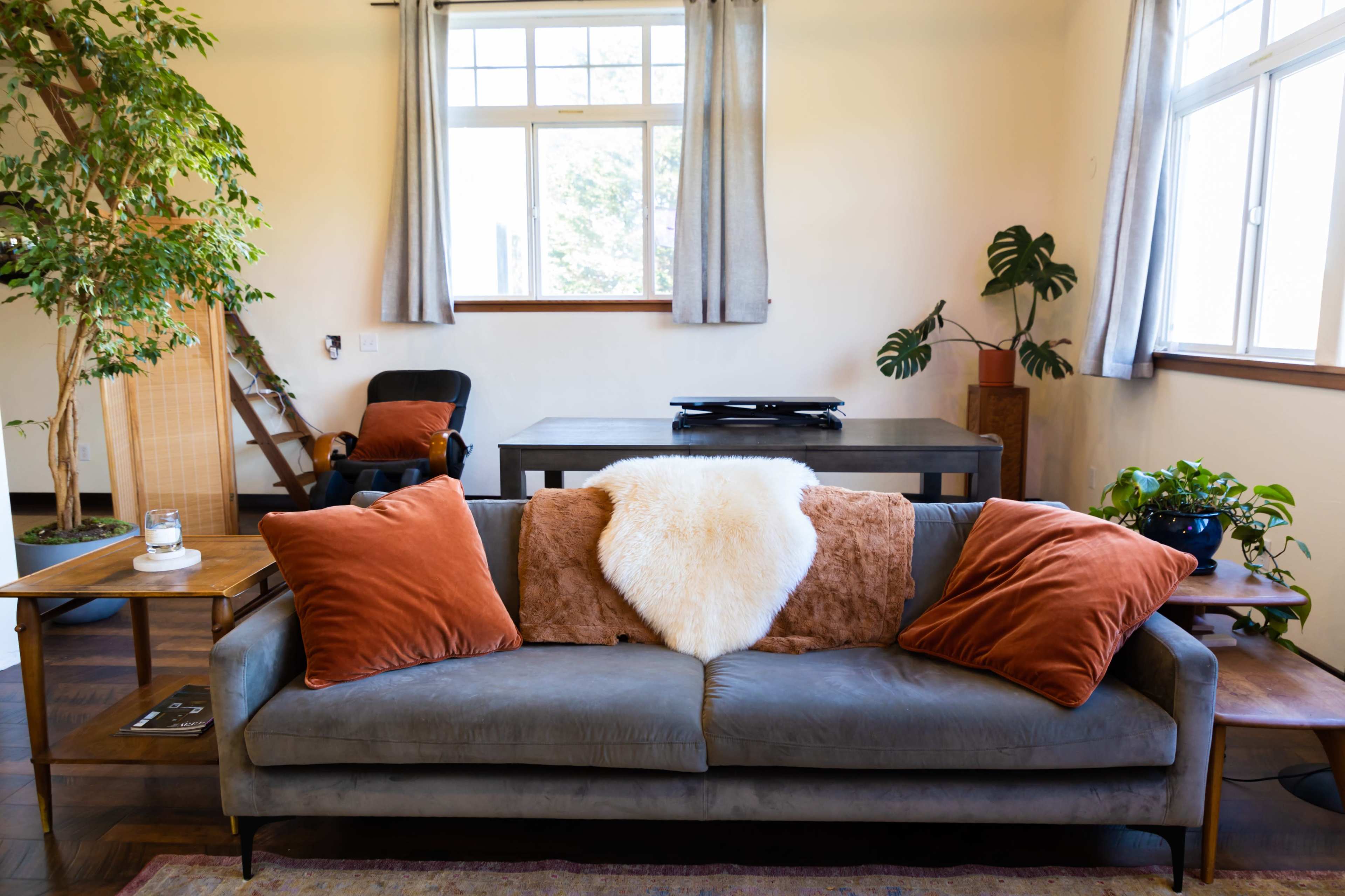 The living room features a gray sofa with orange and brown pillows, a white throw, and surrounding plants near a wooden table and large windows.