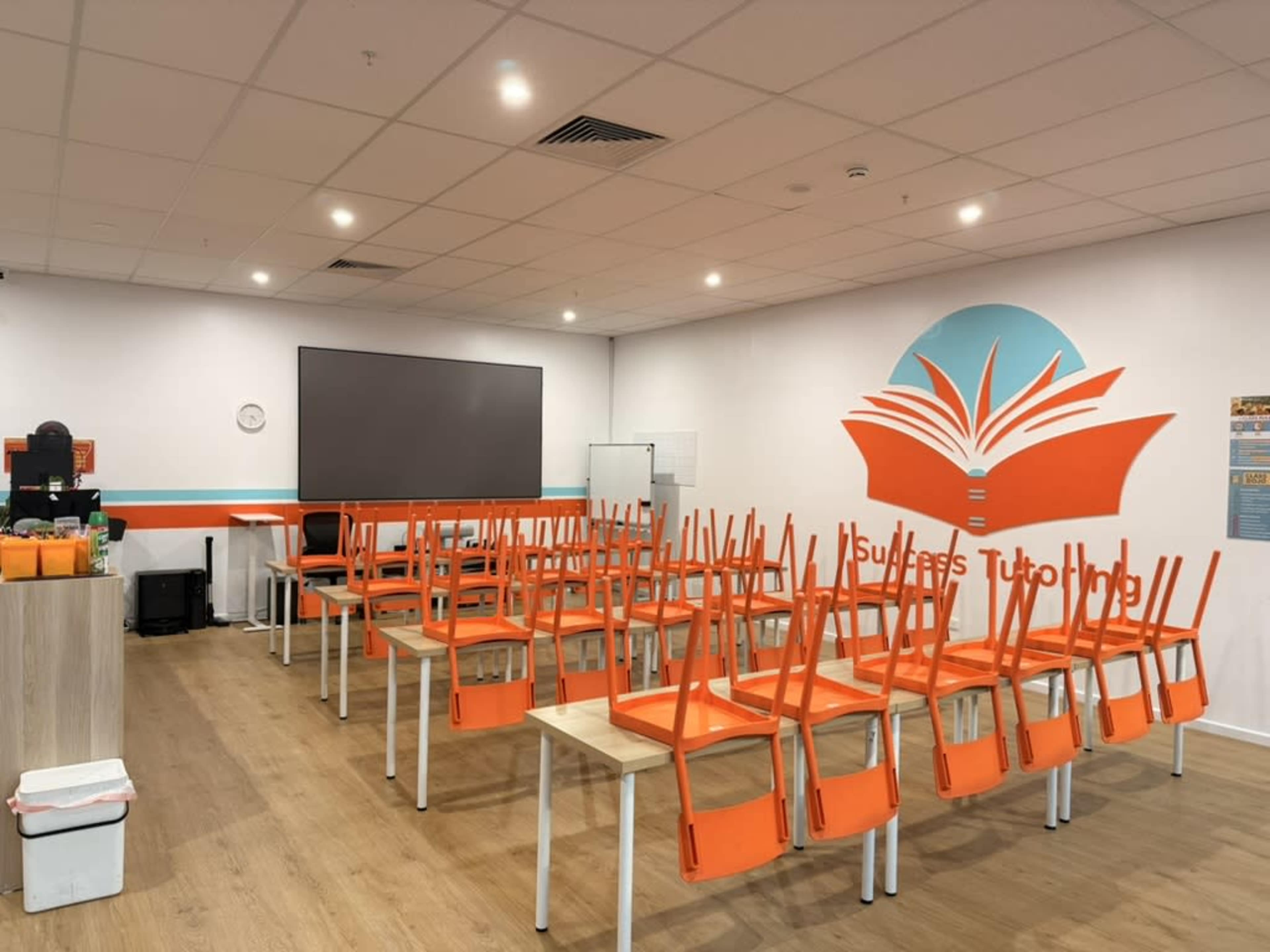 The image shows a tutorial room with tables and chairs stacked upside down, featuring a large wall mural of an open book and bright orange accents.