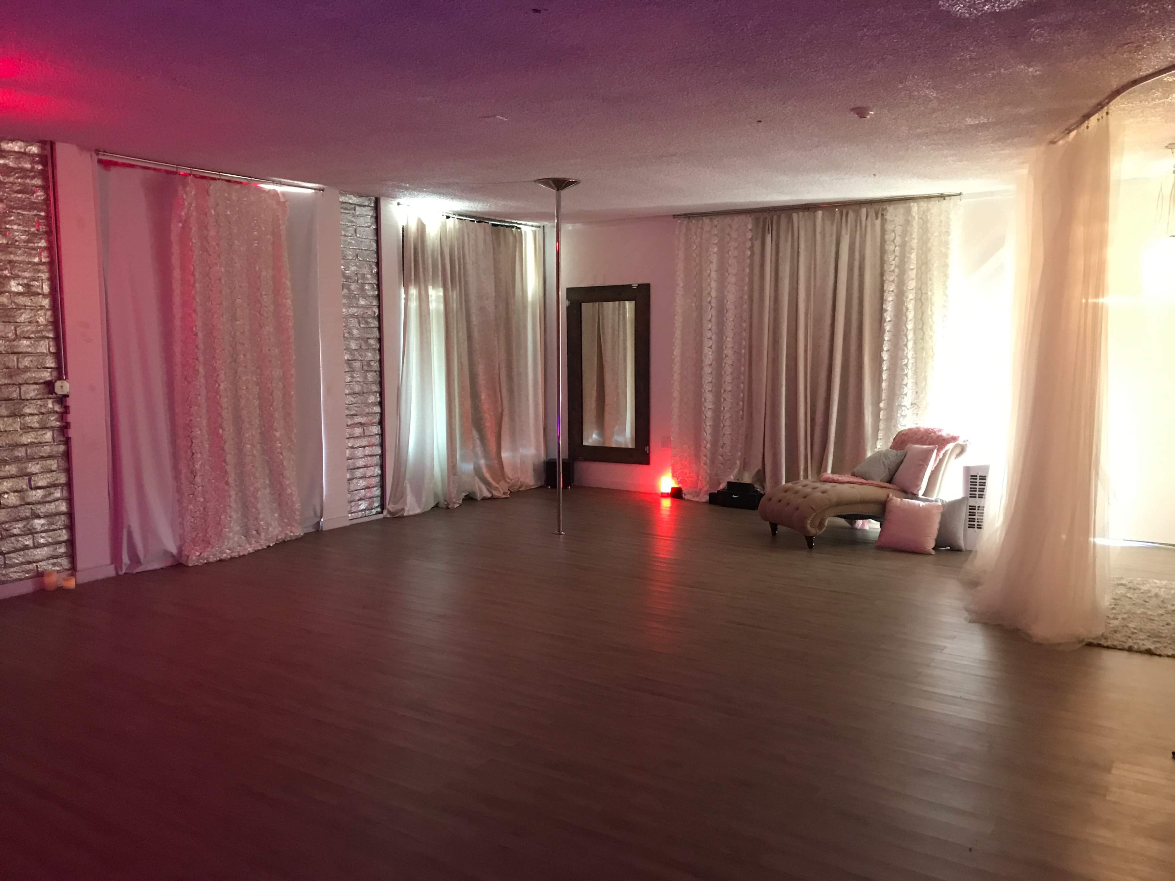 A spacious room with wooden flooring, adorned with sheer curtains, a large mirror, and a pink upholstered chair, illuminated by soft lighting.