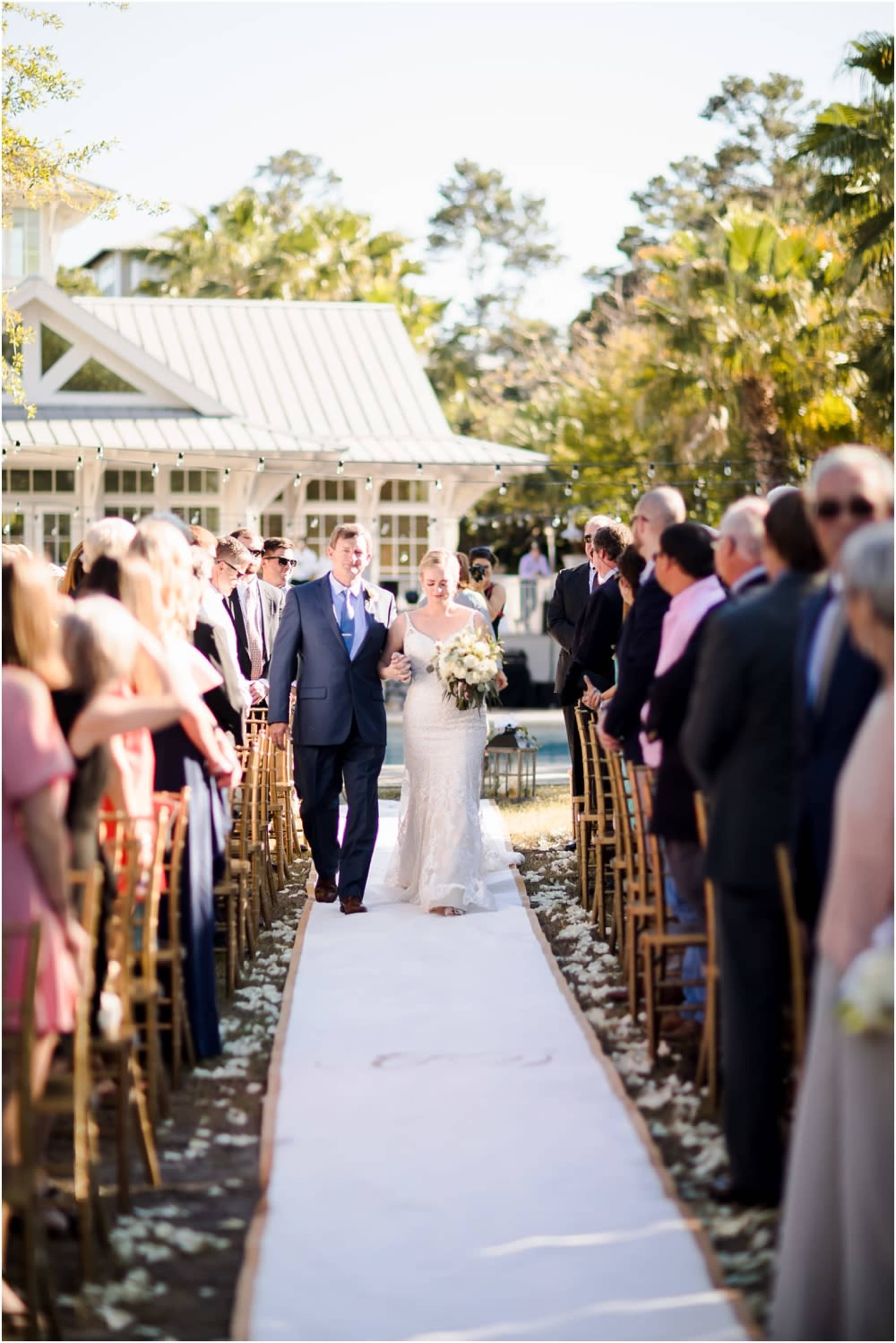 A bride and groom walk down a floral-decorated aisle surrounded by seated guests at an outdoor wedding ceremony.