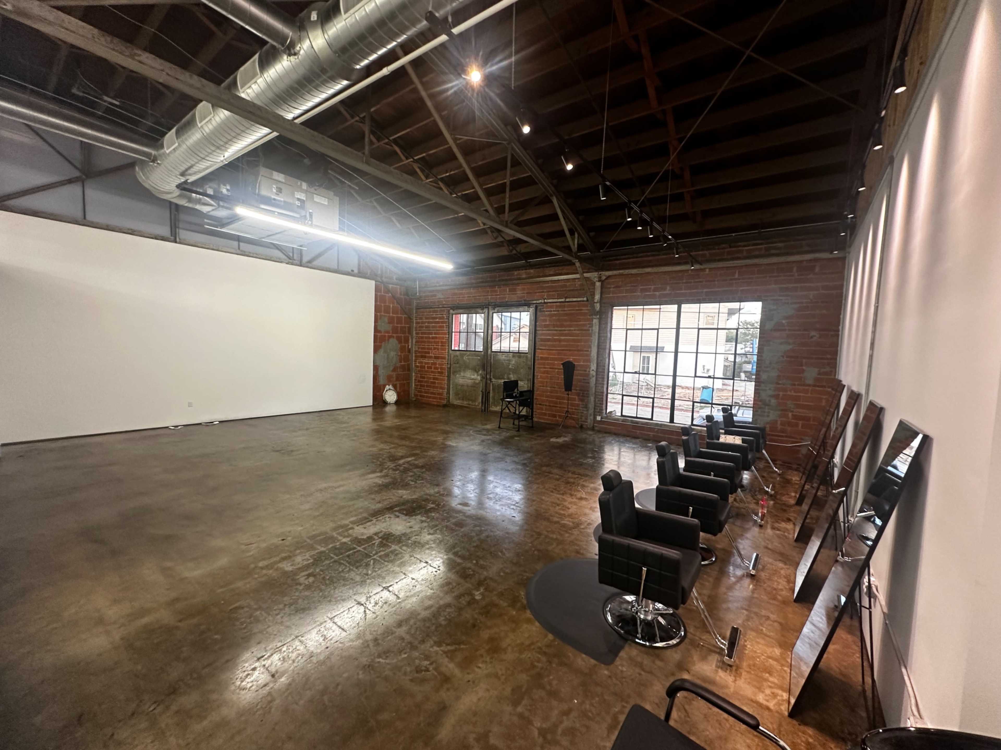 An open, spacious studio with a polished concrete floor, exposed brick walls, and several black salon chairs lined up against the wall.