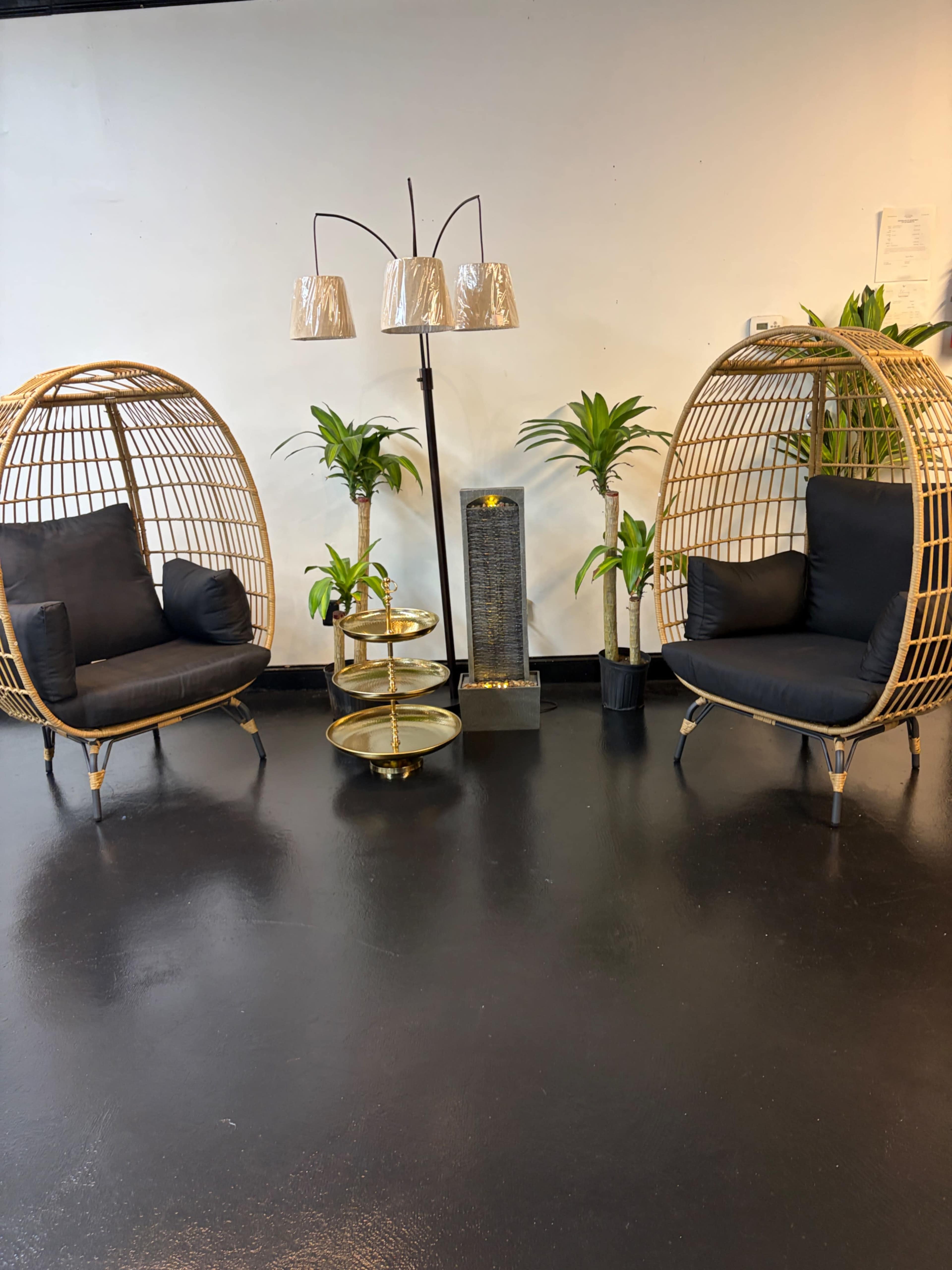 The image shows two rattan chairs with cushions facing each other, surrounded by potted plants and a standing lamp with three shades.
