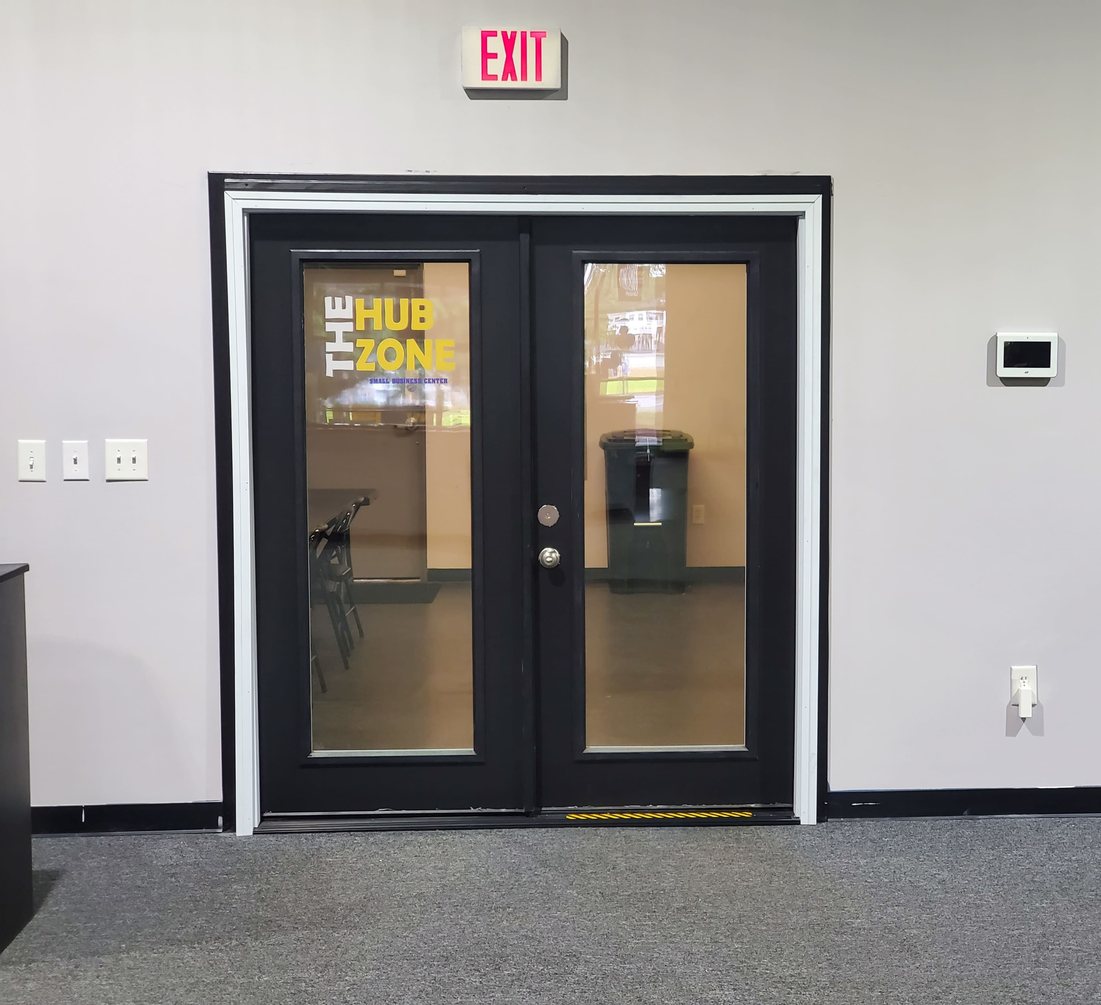 The image shows a set of double glass doors labeled "THE HUB ZONE," with an exit sign above and a visible room behind them.