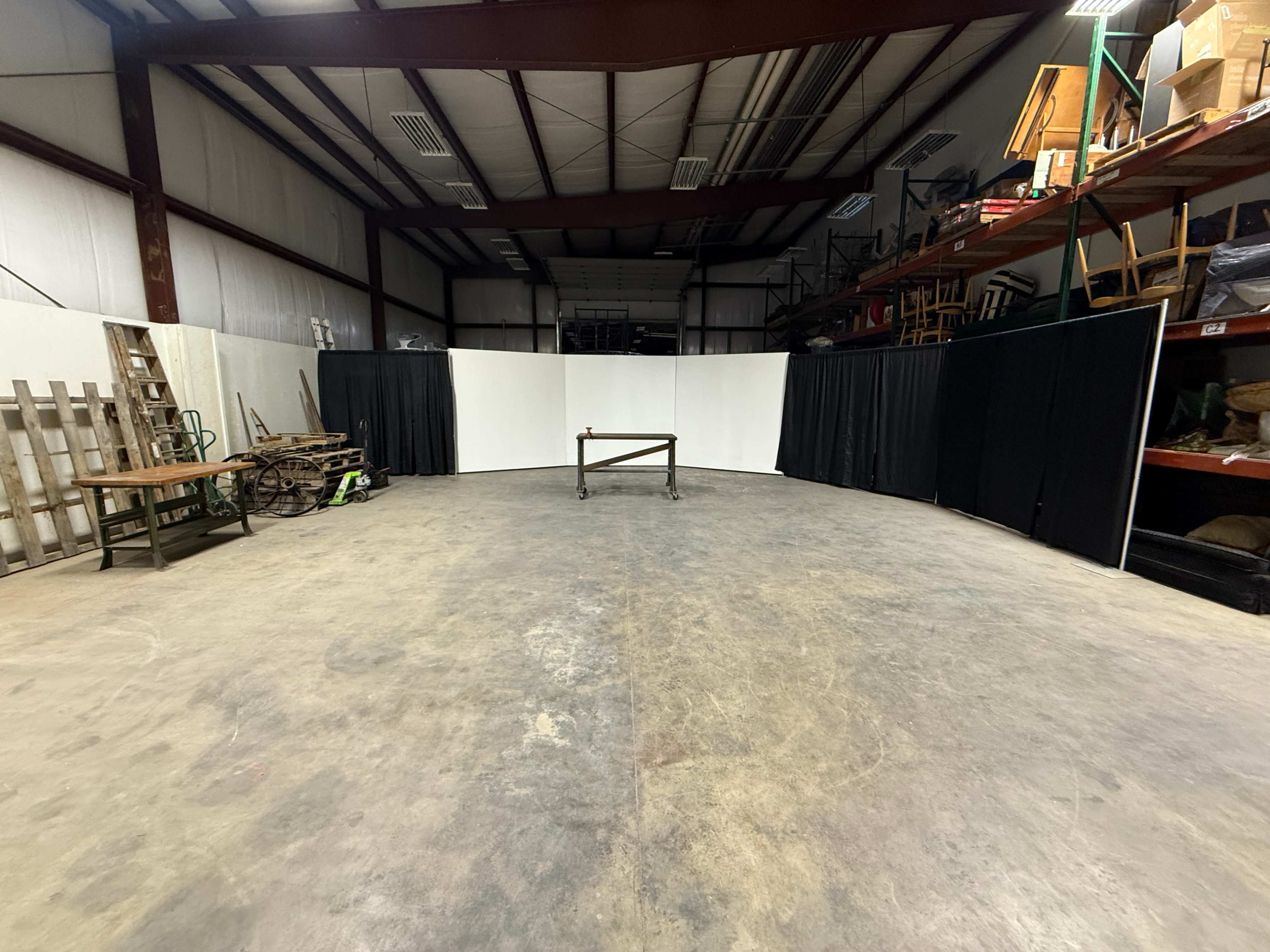 The image shows a large industrial space with a curved arrangement of plain white and black curtains, surrounded by shelves and various equipment.