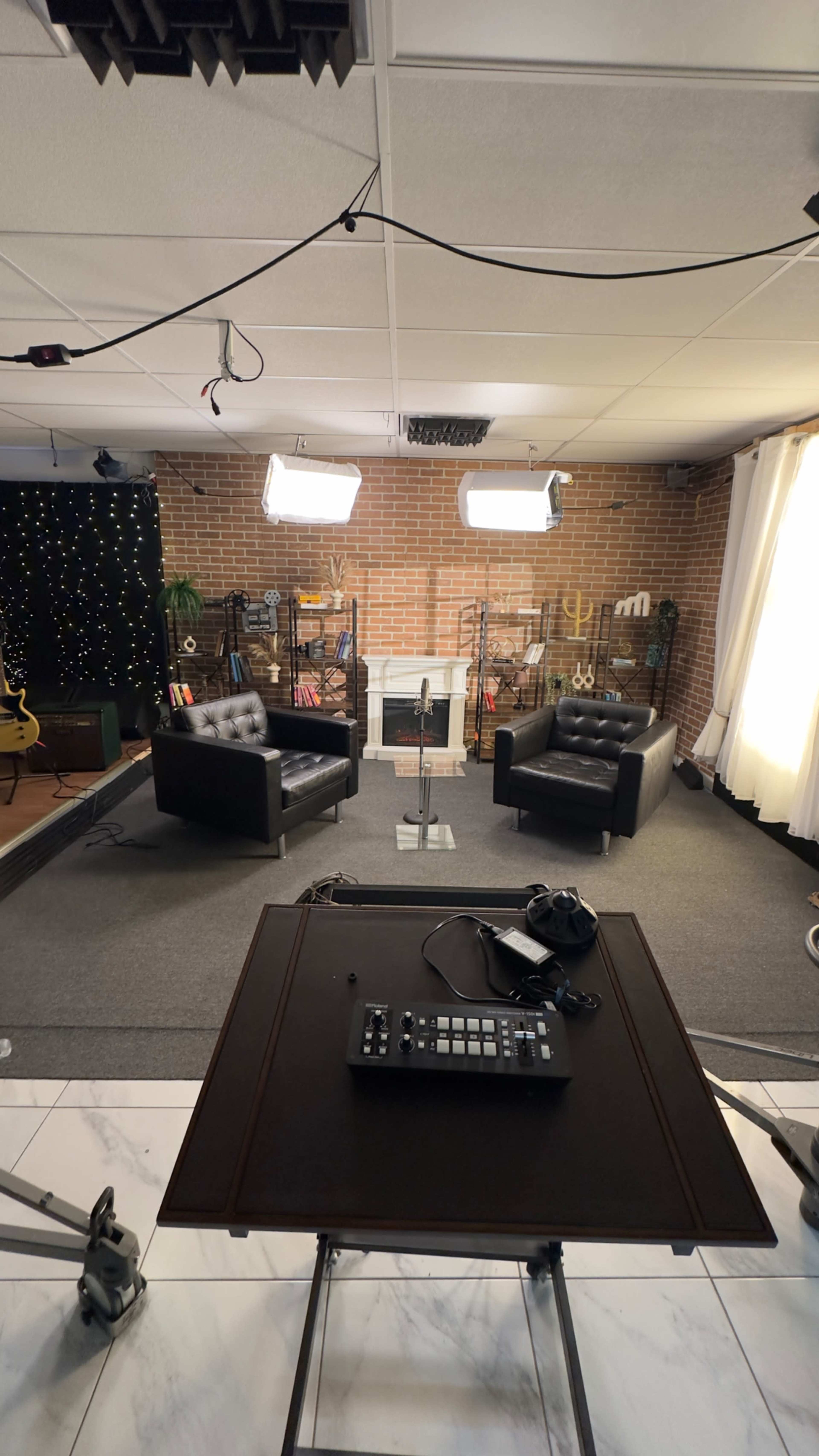 The image shows a modern studio setup featuring two black leather chairs facing each other, a microphone stand in the center, and soft lighting against a brick wall with decorative elements.