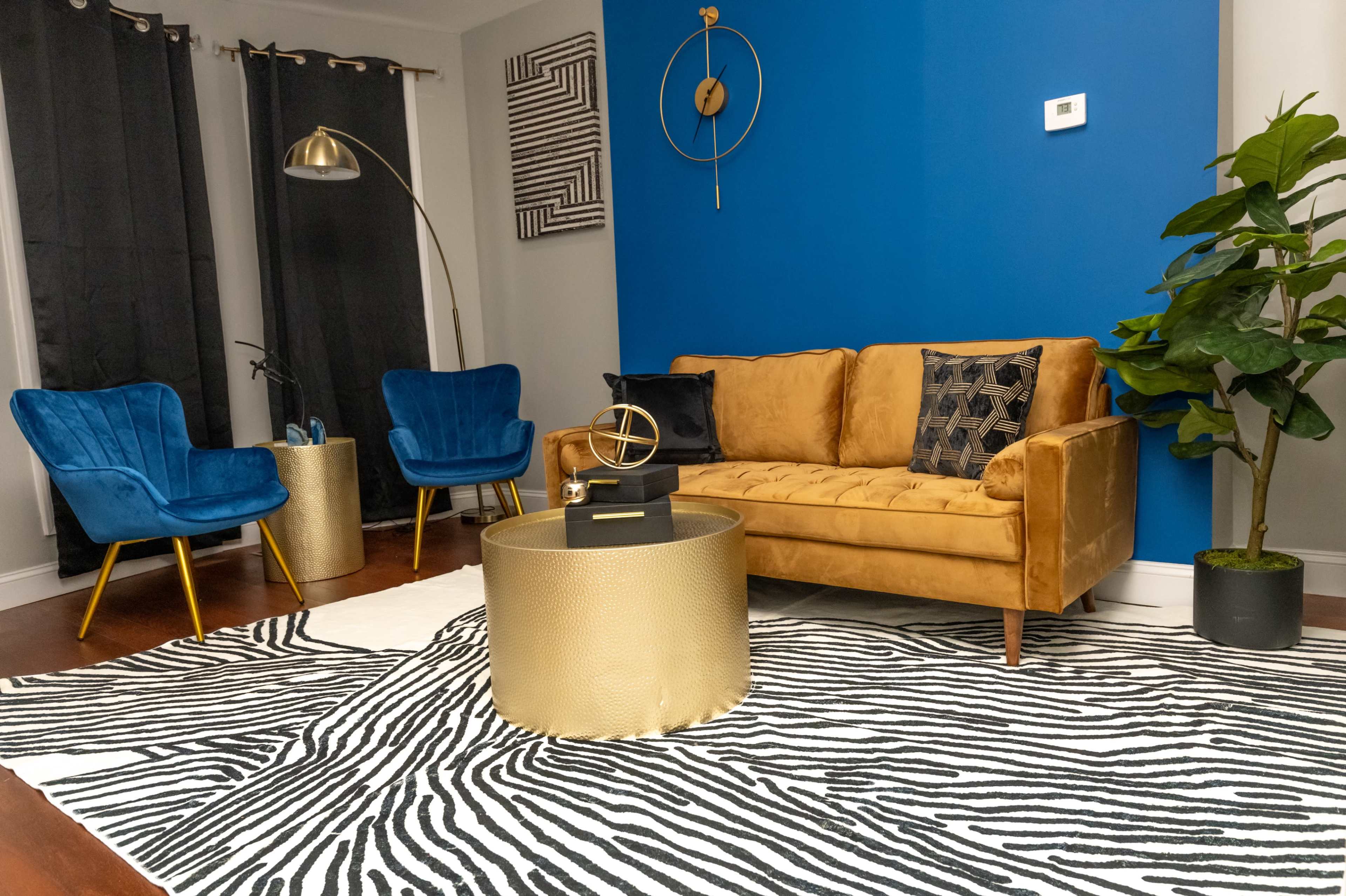 A living room with a golden sofa, two blue armchairs, a zebra-striped rug, a gold coffee table, and decorative plants against a blue accent wall.