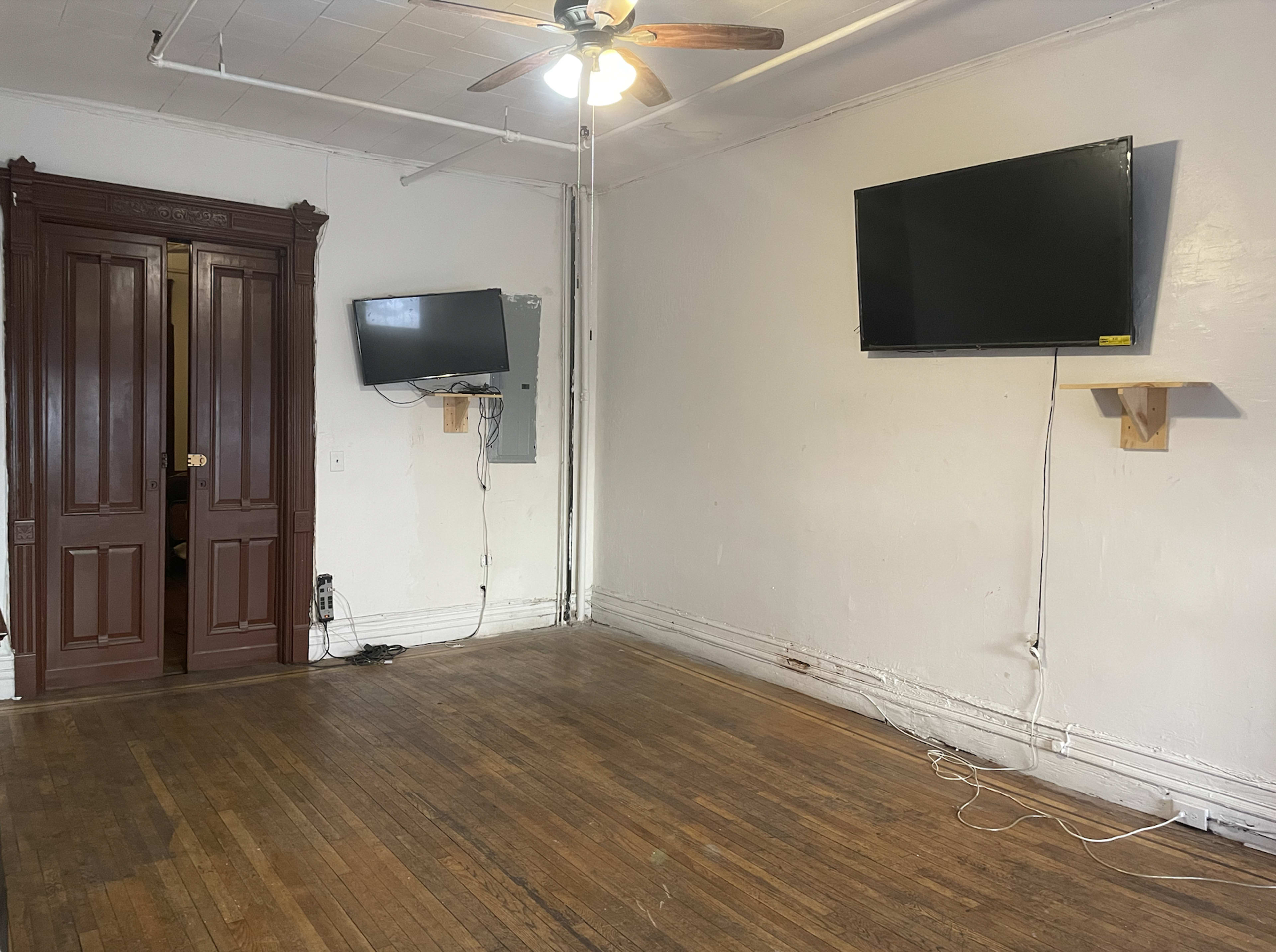 The image shows a room with hardwood flooring, two mounted televisions on the walls, and a set of double doors on one side.