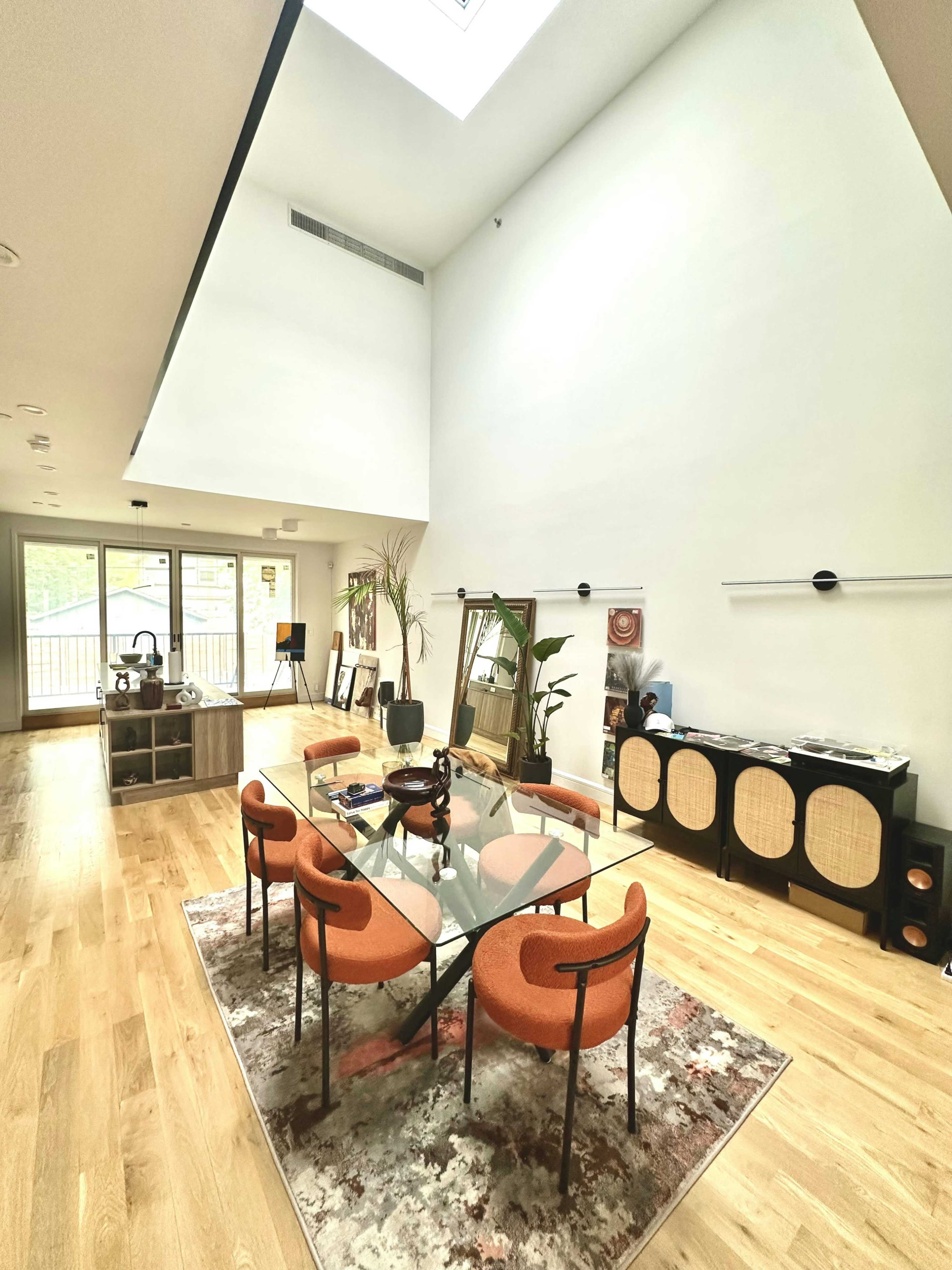 The image shows a spacious, well-lit modern living area with a glass dining table surrounded by orange chairs, a wooden cabinet, and large windows.