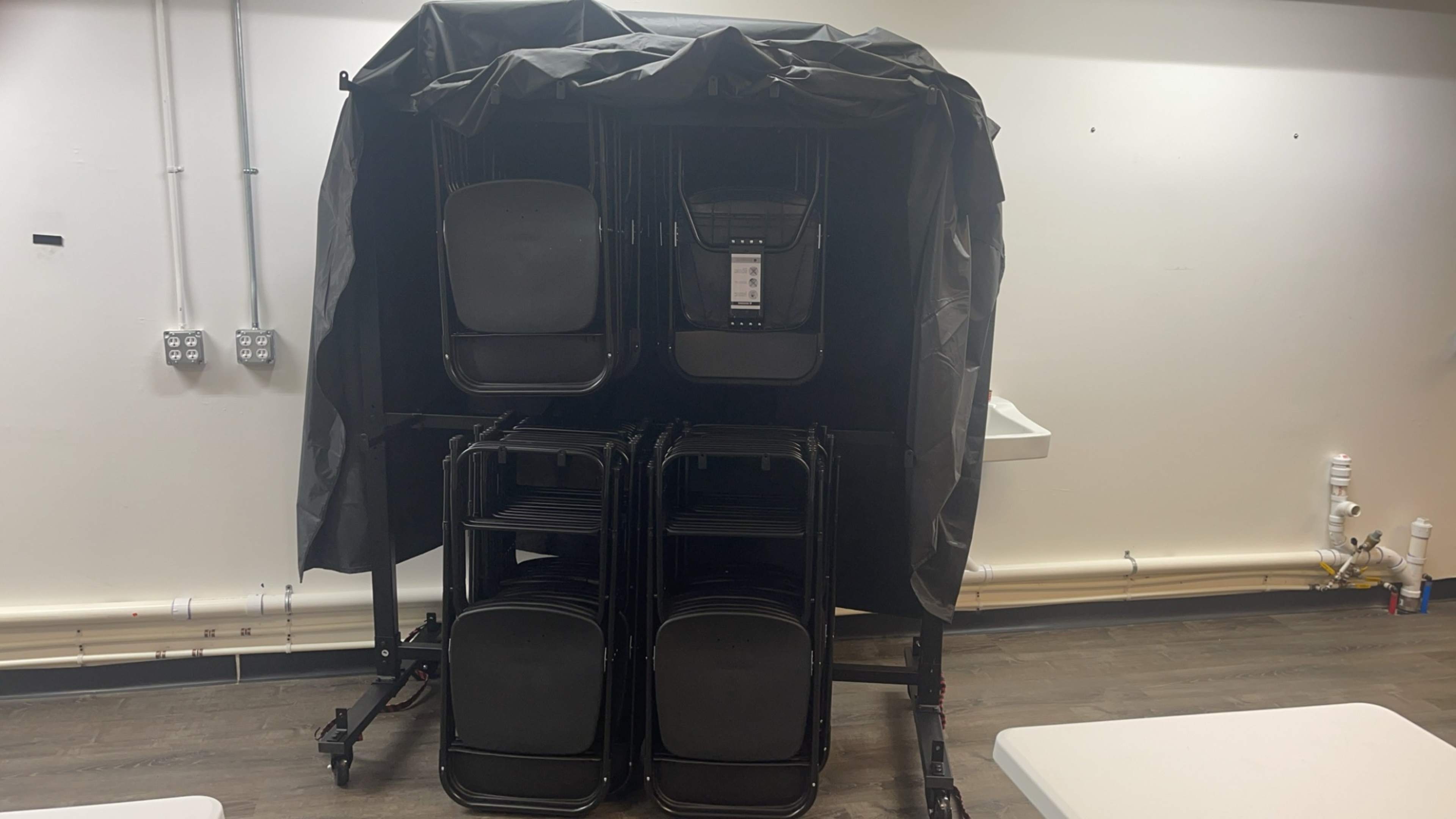 A stack of black chairs is covered with a black tarp and secured against a wall in a room.