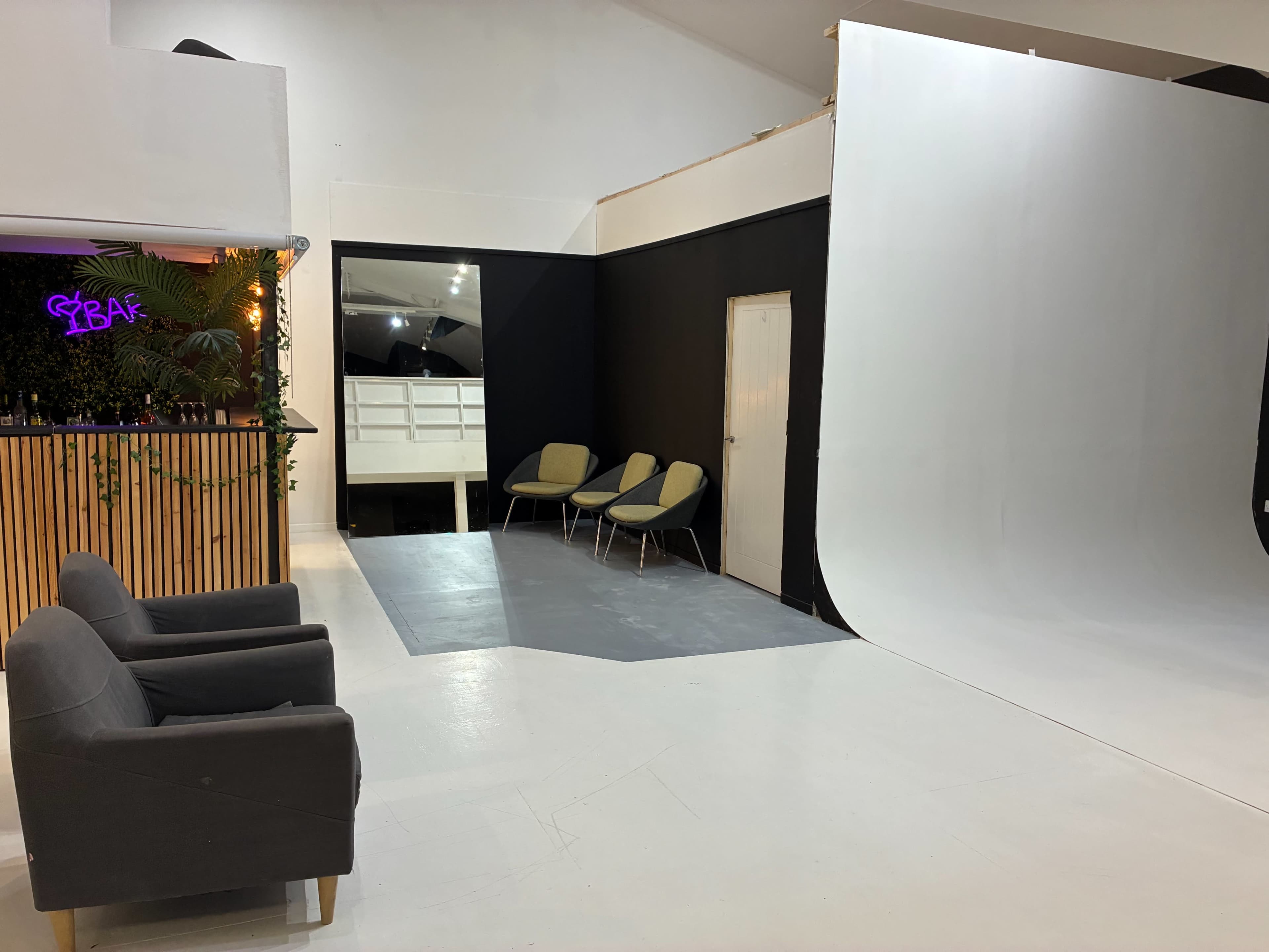 The image shows a photography studio featuring a white backdrop, two gray chairs, and a small seating area with green chairs against a black wall.