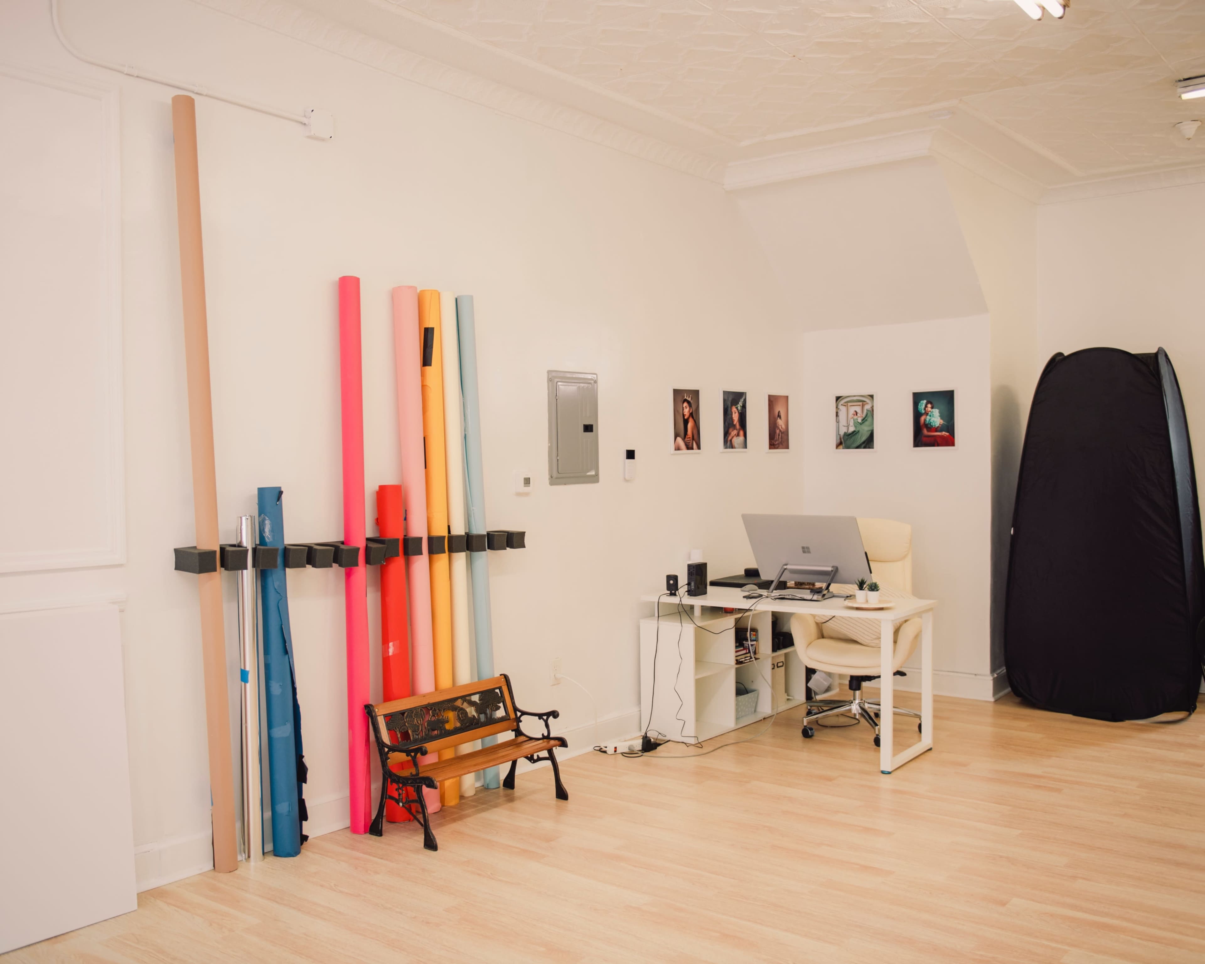 A minimalistic studio space with colorful rolls of material on the left, a desk with a computer set up in the center, and a portable backdrop in the corner.