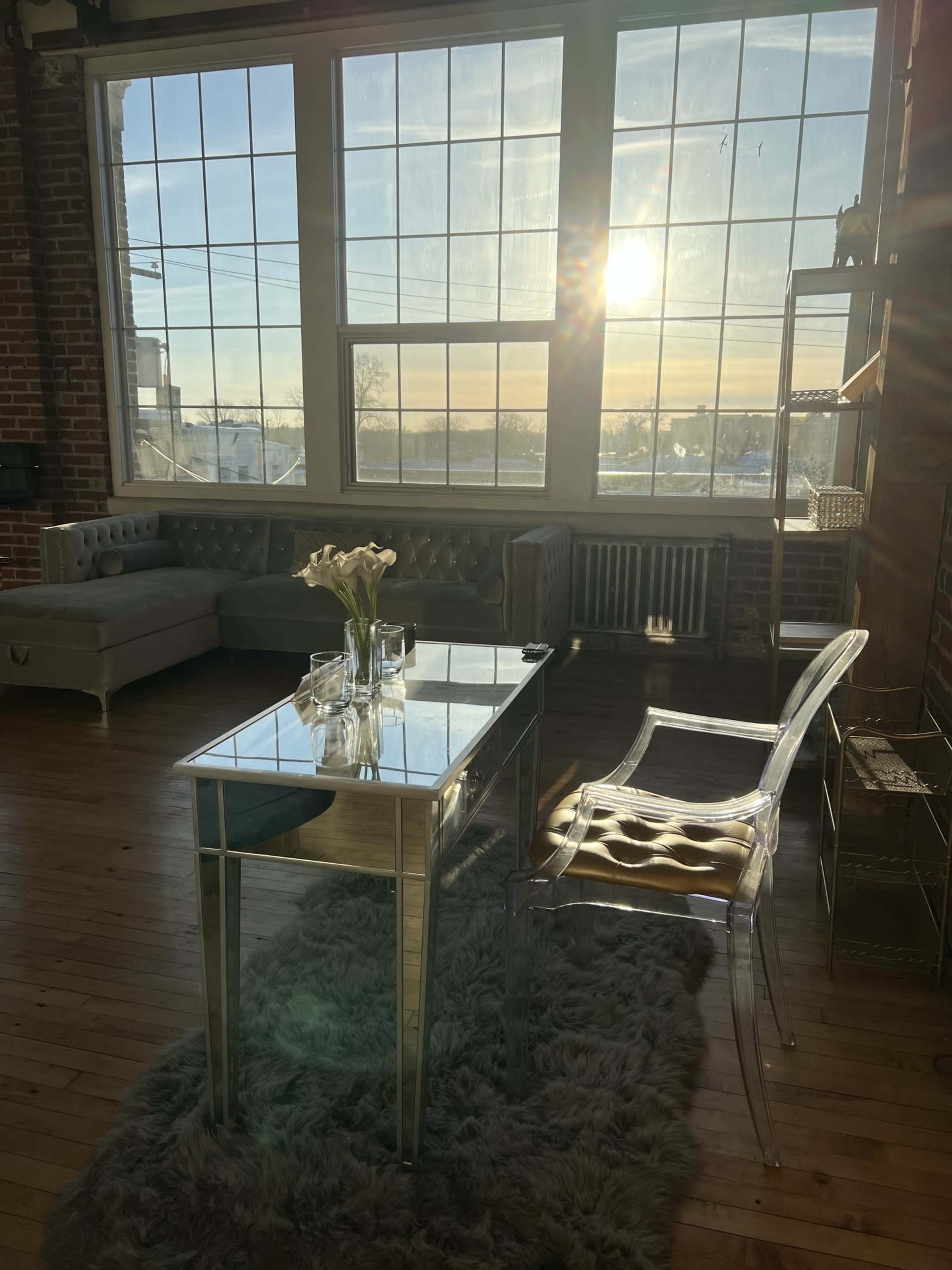 A modern office space features a glass-top table, a transparent chair, and a bouquet of flowers, with sunlight streaming through large windows.