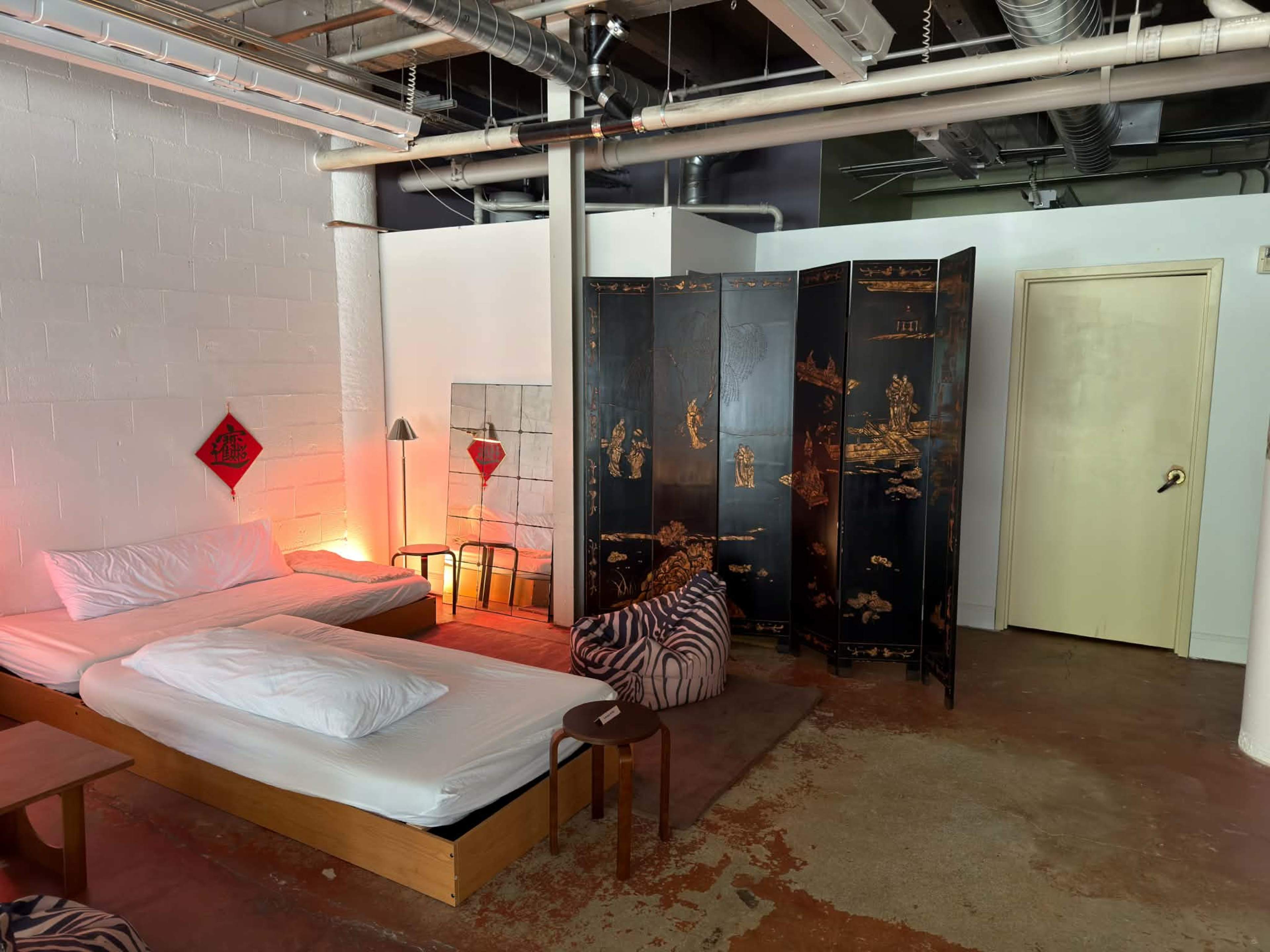 The image features a minimalist bedroom with a wooden bed, a folding screen, and a seating area, all set in a space with exposed pipes and industrial elements.