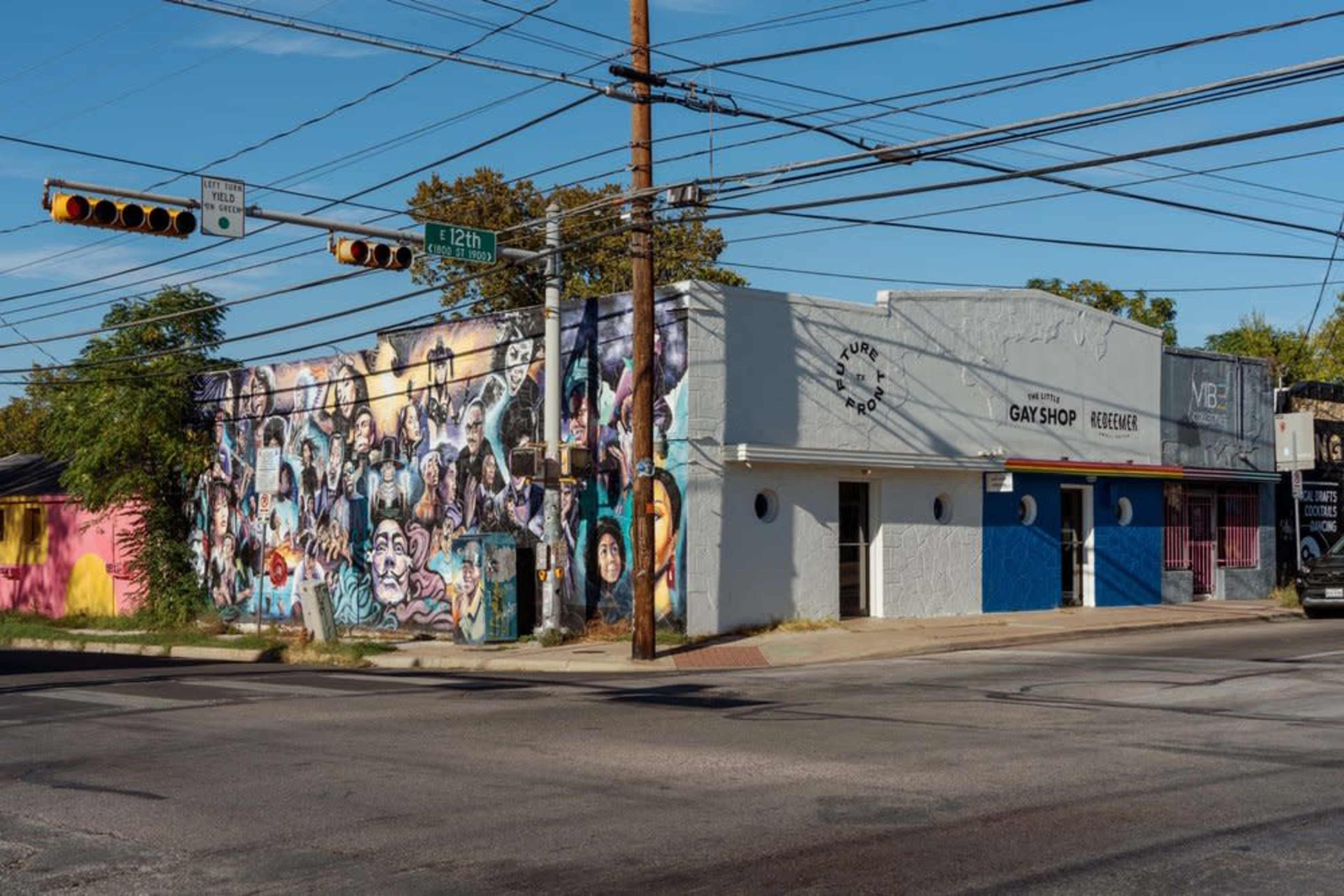 A street corner featuring a building with a colorful mural on one side, a gay shop, and a blue storefront.