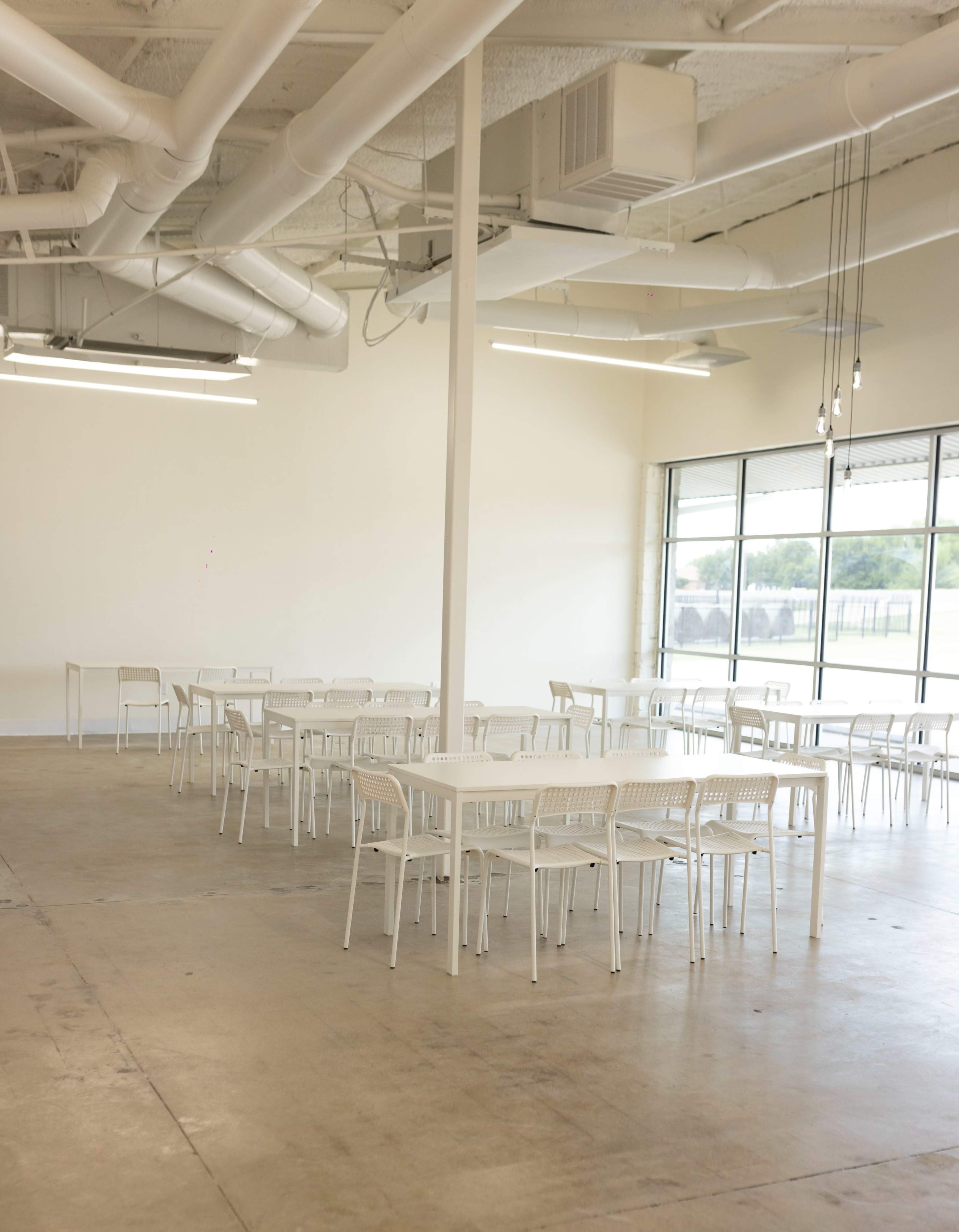 The image shows a spacious, empty dining area with several white tables and chairs arranged neatly, and large windows allowing natural light to enter.