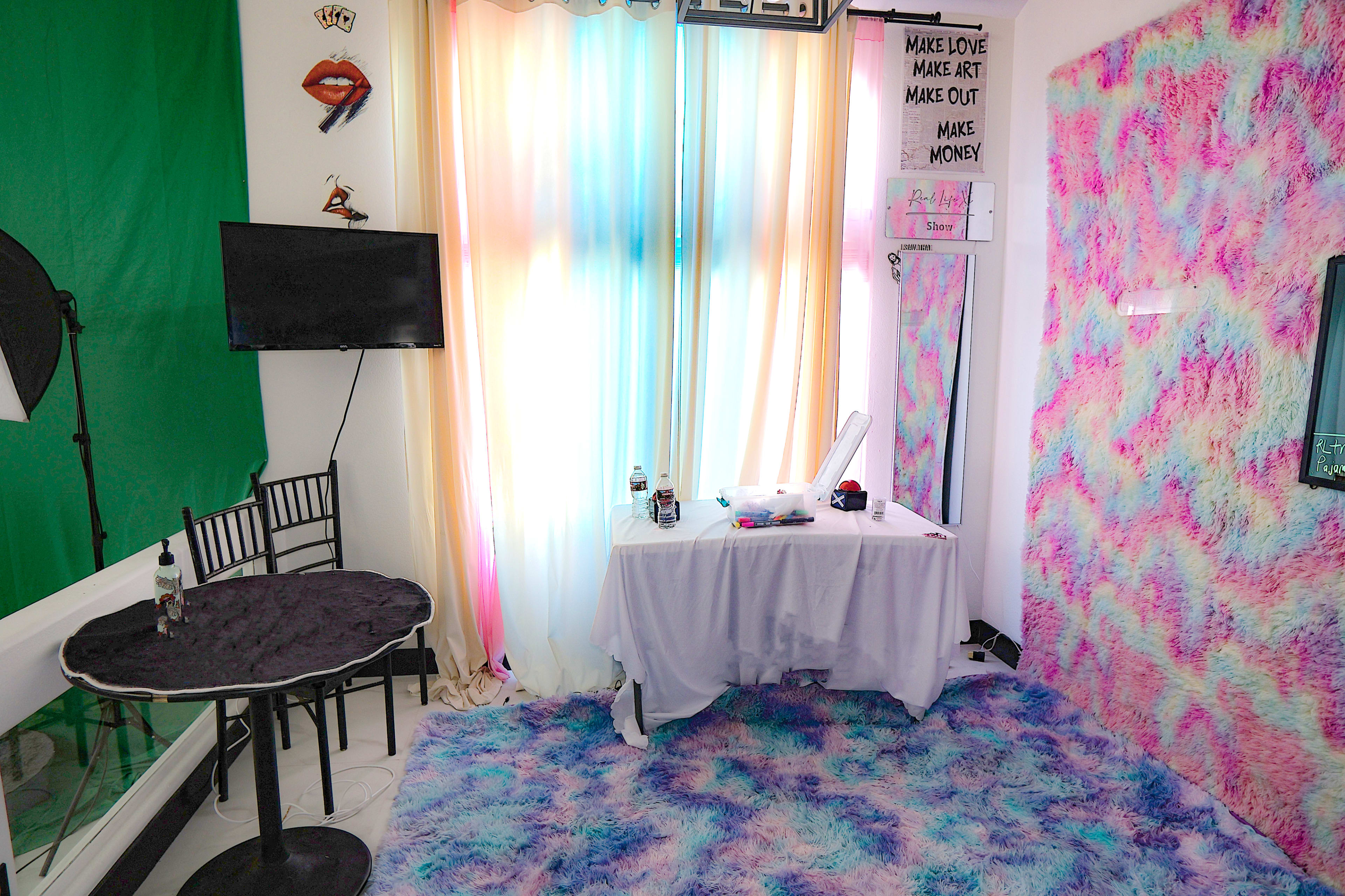 The image shows a small room with colorful curtains, a table covered with a white cloth, and walls adorned with a patterned textile, featuring a television and a black chair.