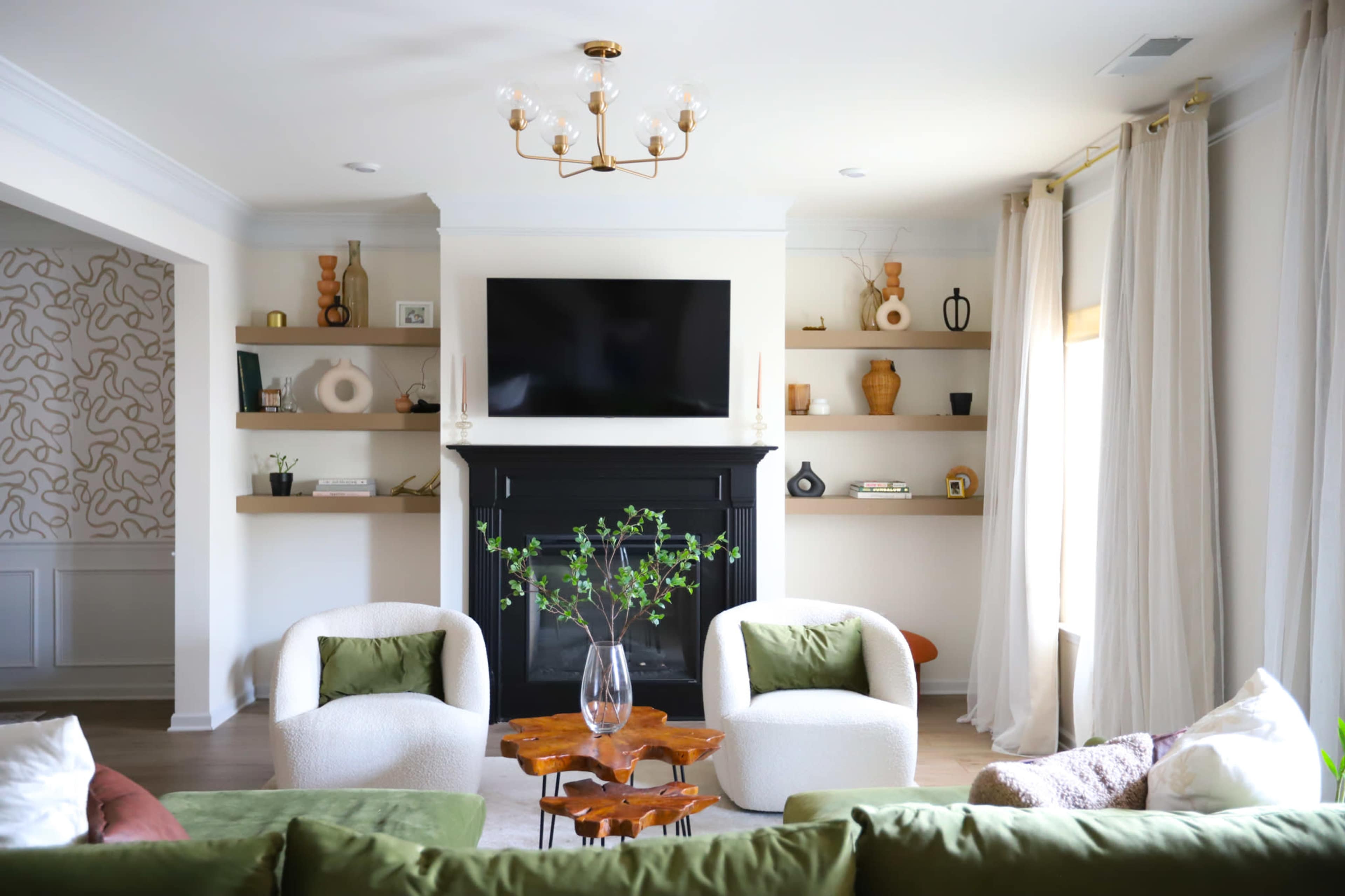 A modern living room features two upholstered chairs, a sofa, a black fireplace, and shelves with decorative items and plants.