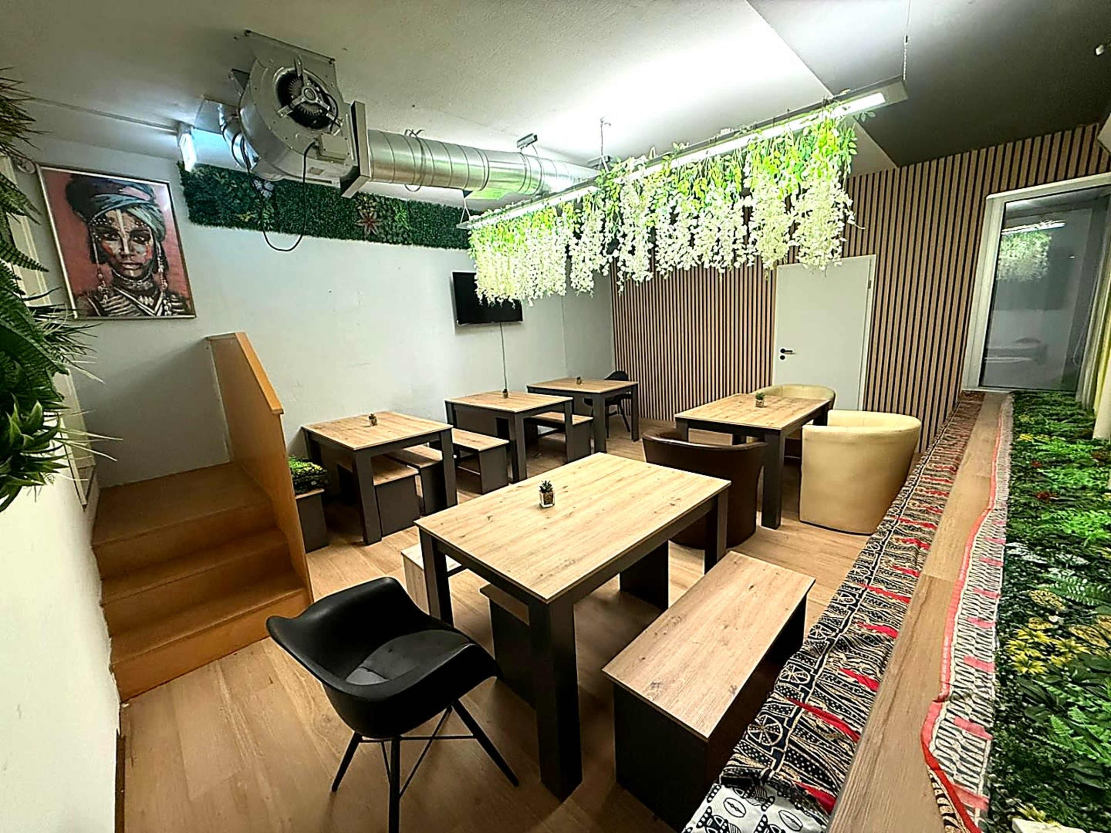 The image shows a modern, minimally decorated café or meeting room featuring wooden tables, benches, and a staircase, with greenery and wall art enhancing the ambiance.