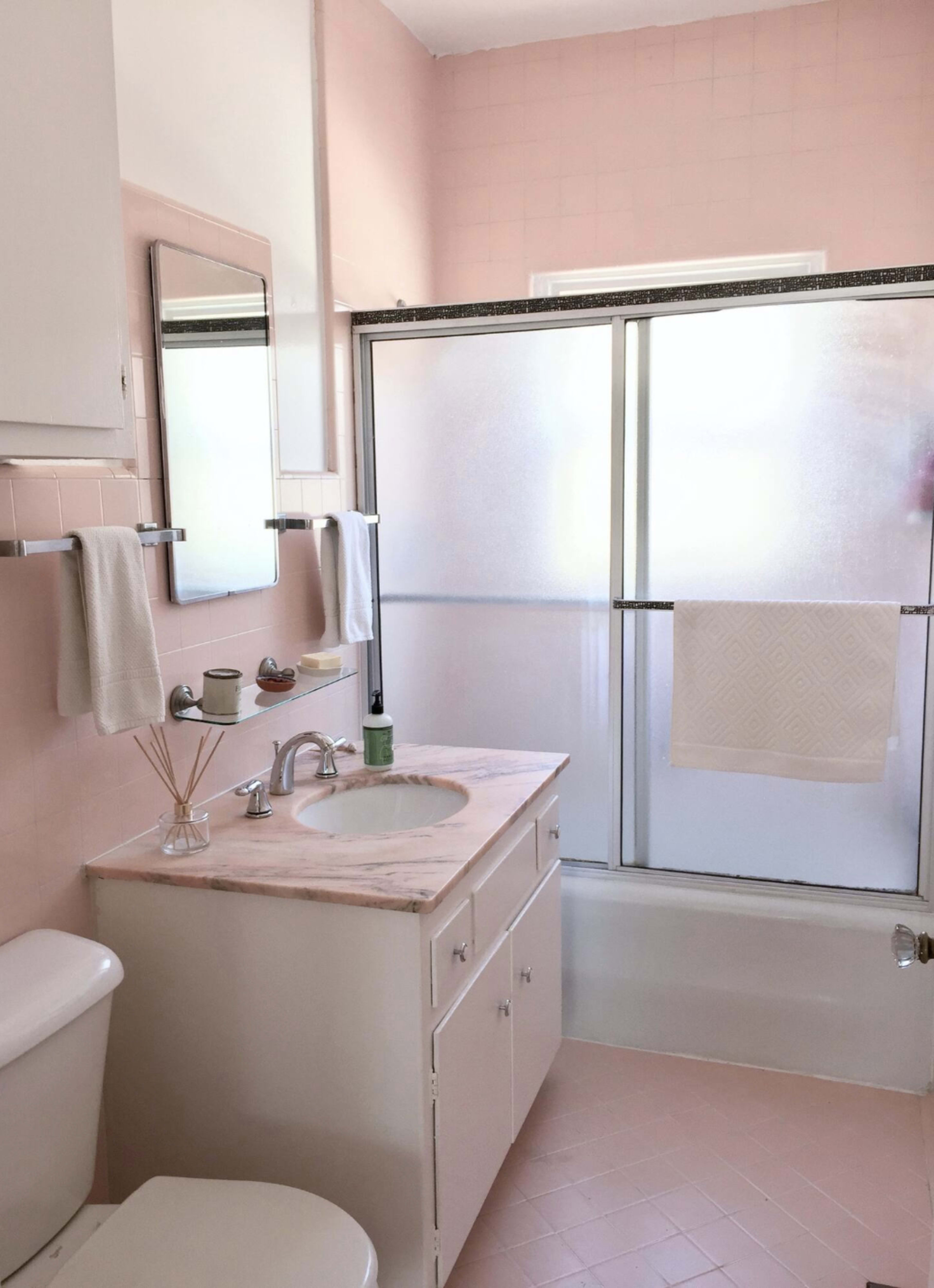 The image shows a light pink bathroom with a white vanity, a mirror above the sink, a shower stall with a sliding glass door, and a tub beneath a window.