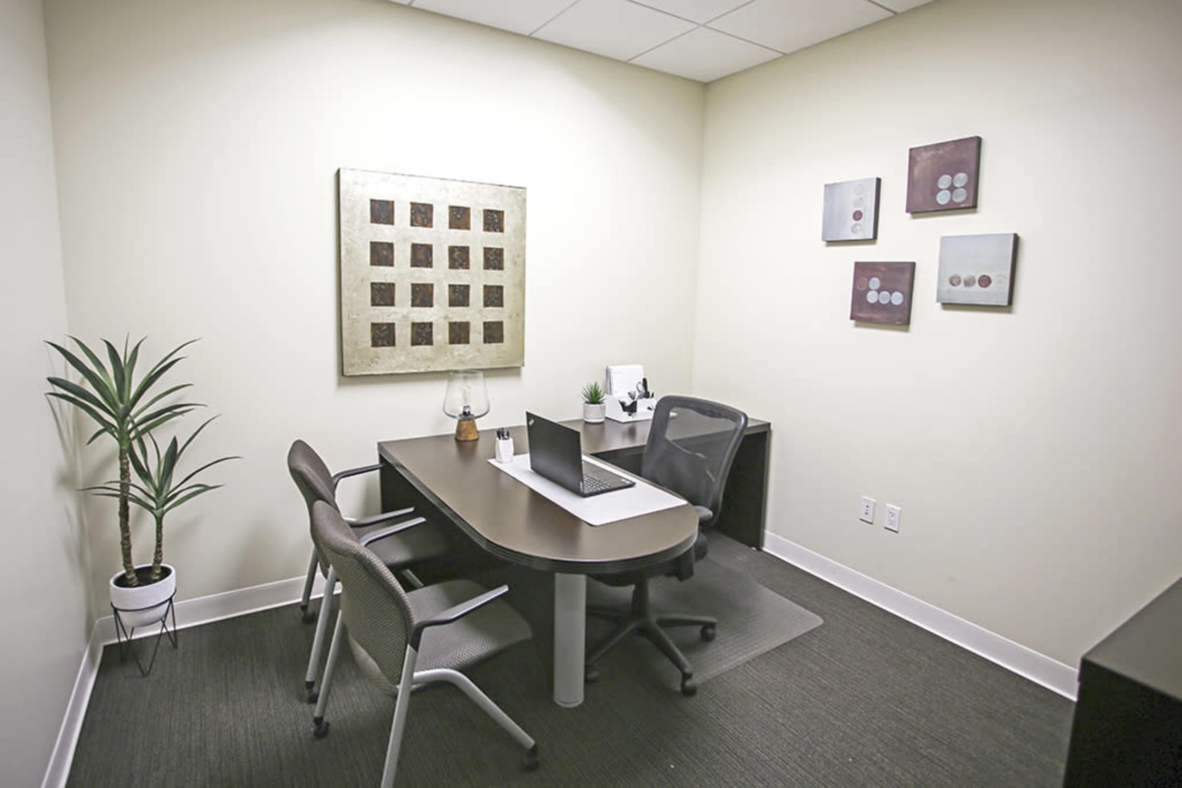 The image shows a small office space featuring a desk with a laptop, two chairs, a plant, and framed wall art.