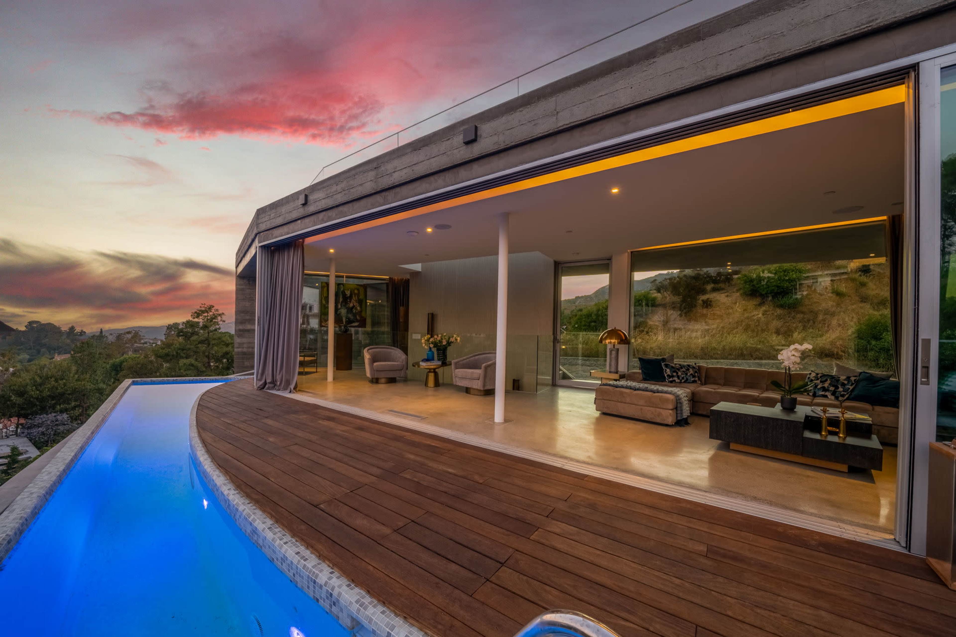 A modern house features an expansive terrace with a swimming pool, overlooking a sunset view with vibrant clouds.