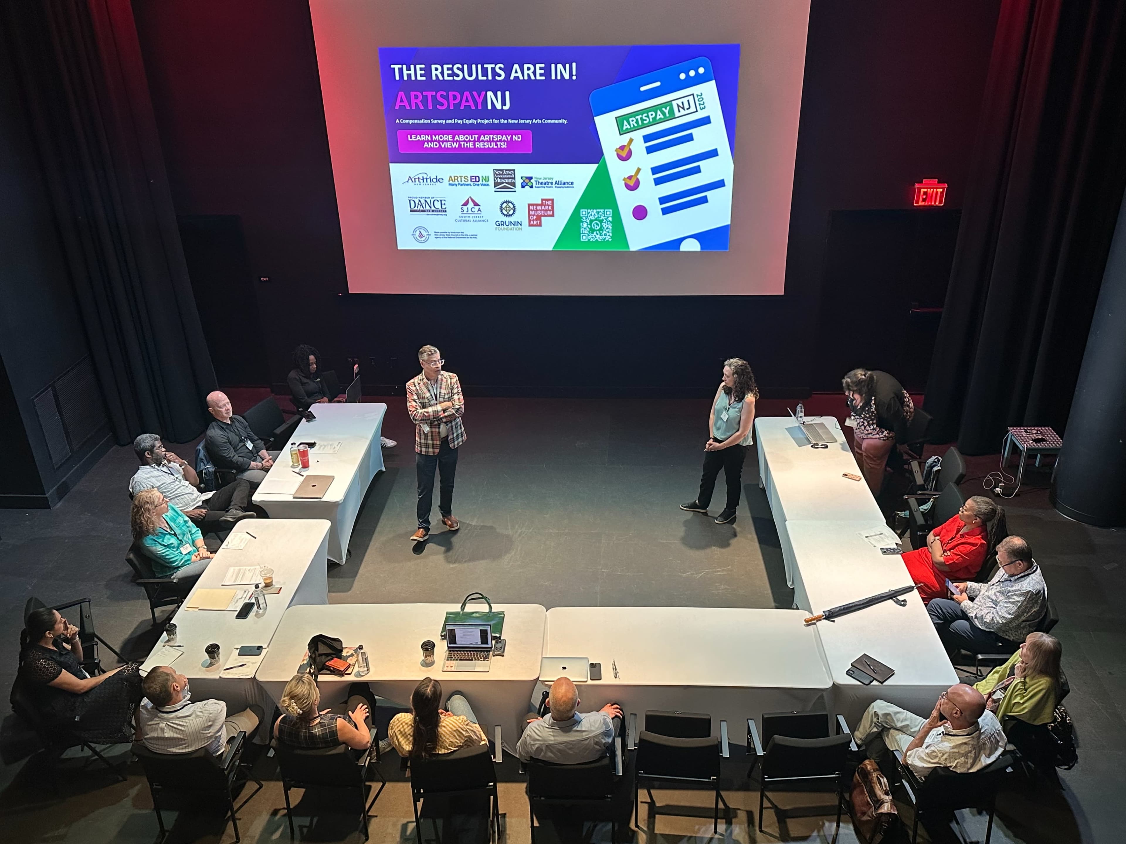 A group of people is seated around a rectangular space in a dimly lit room, while two presenters stand in front of a large screen displaying the text "THE RESULTS ARE IN! ARTSPAY NJ."