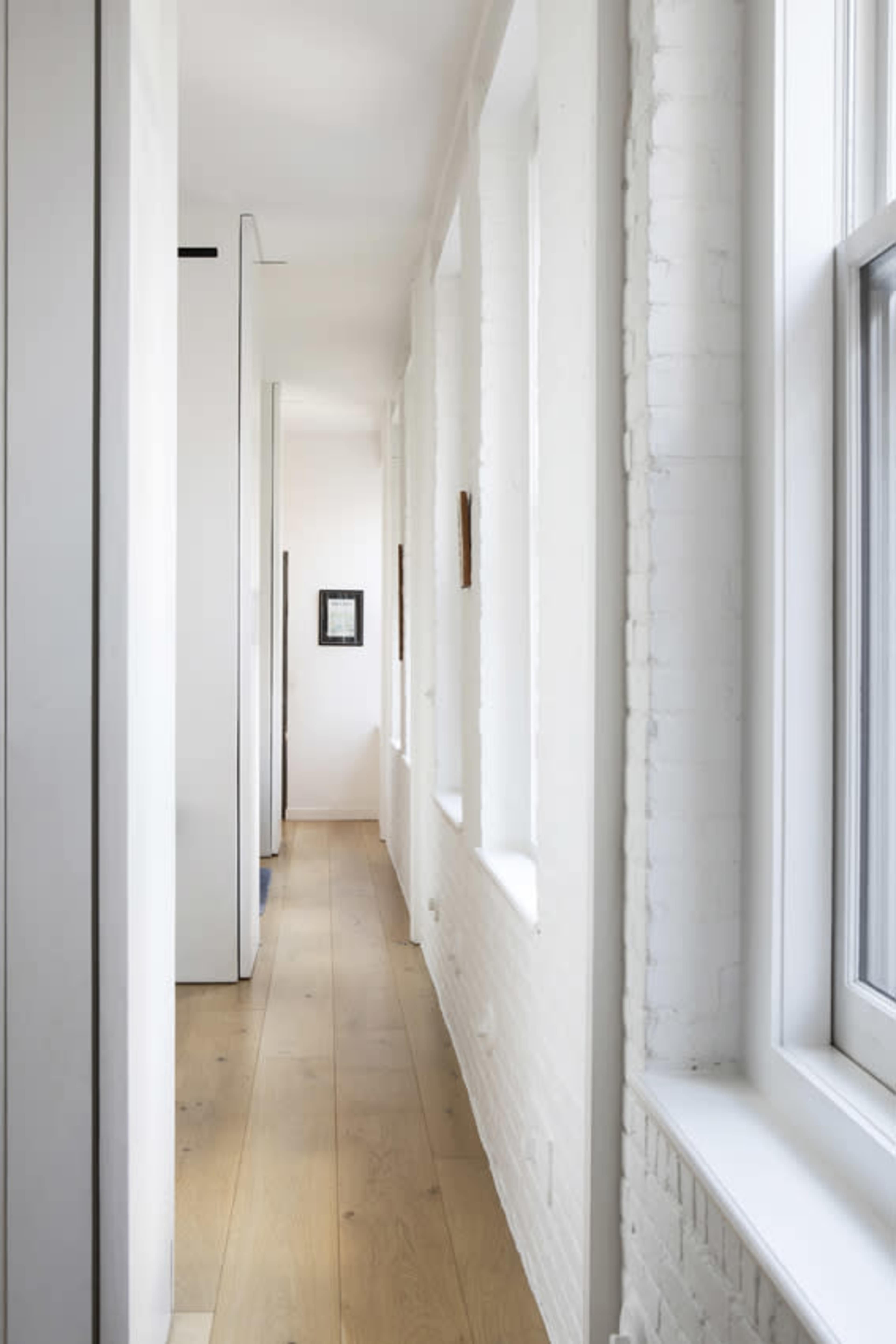 A narrow, bright corridor features white brick walls and large windows with wooden flooring.