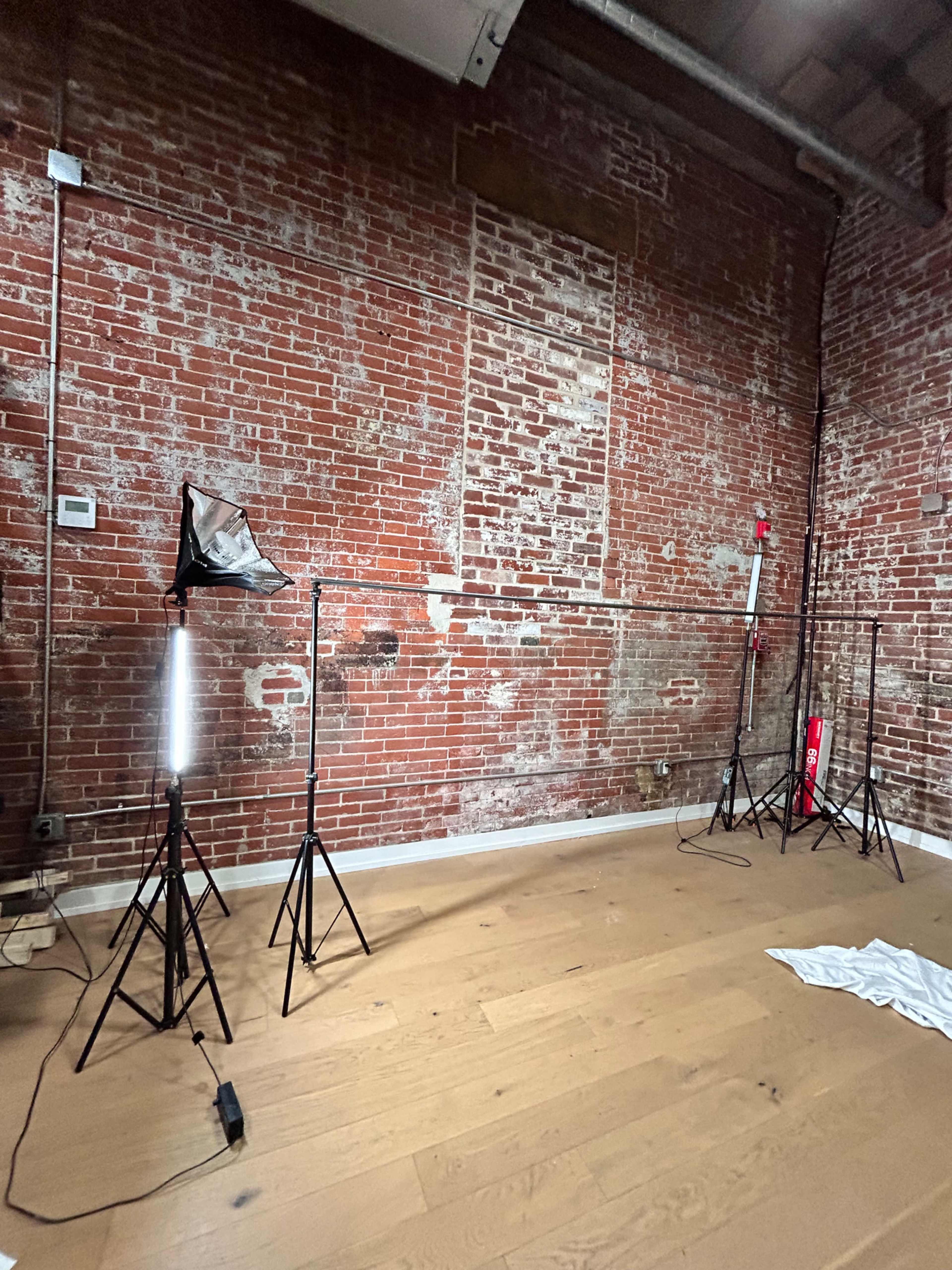 The image shows a room with a wooden floor and exposed brick walls, featuring several tripod light stands positioned around the space.