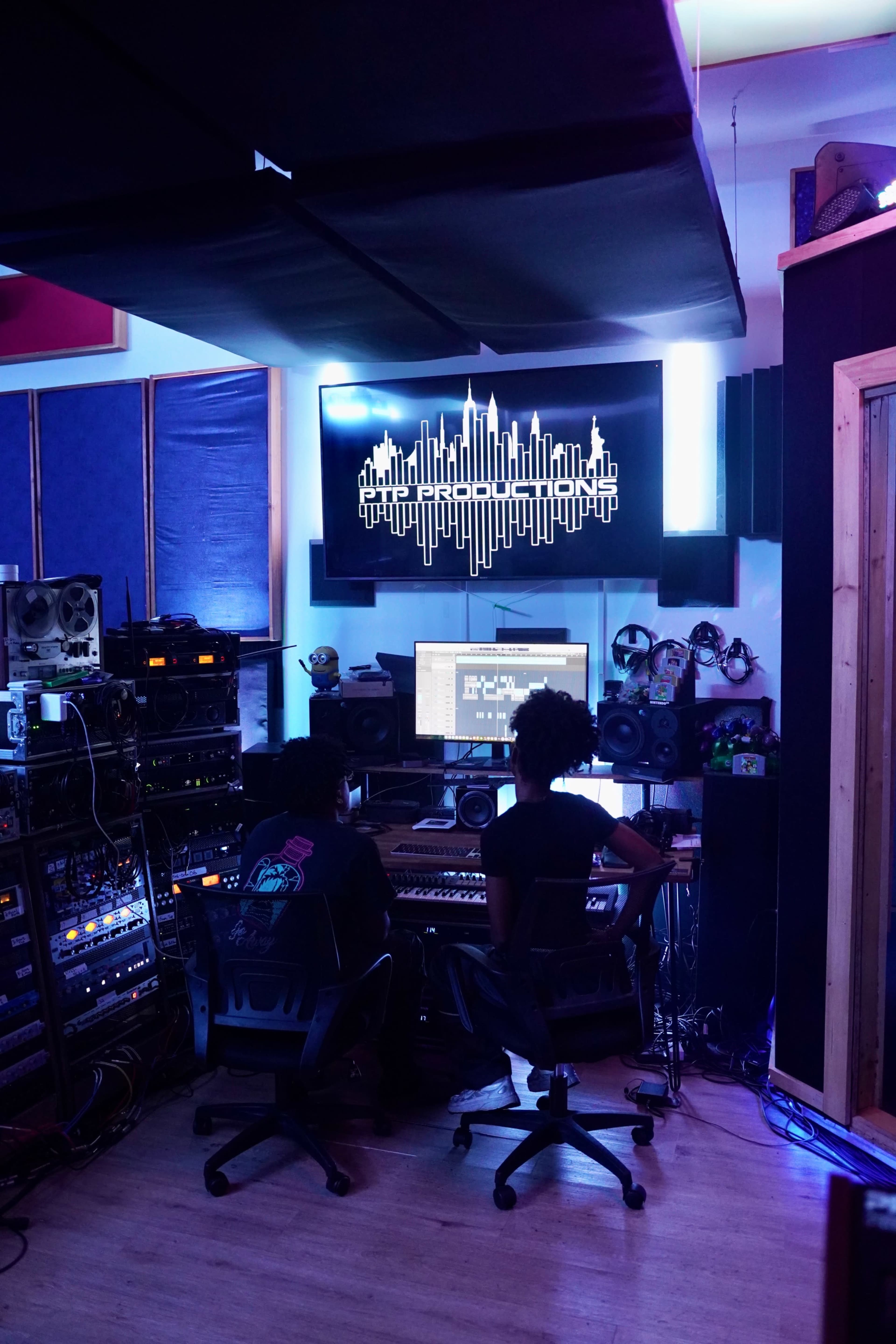 Two individuals work on a music production project at a recording studio, seated in front of a computer with audio equipment and sound panels surrounding them.