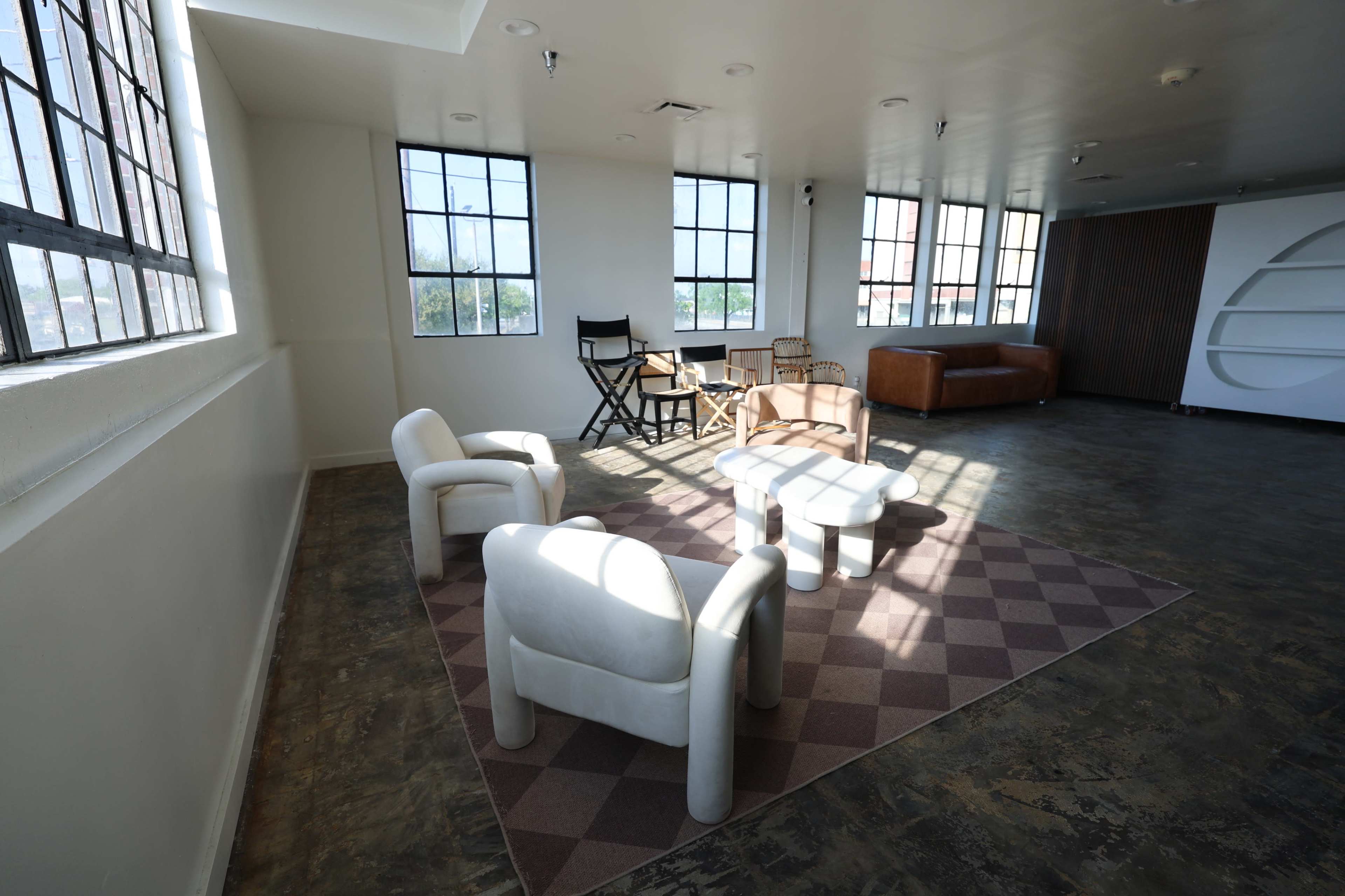 The image shows a modern, open room with large windows, featuring a few chairs, a small table, and a brown couch on a patterned rug.