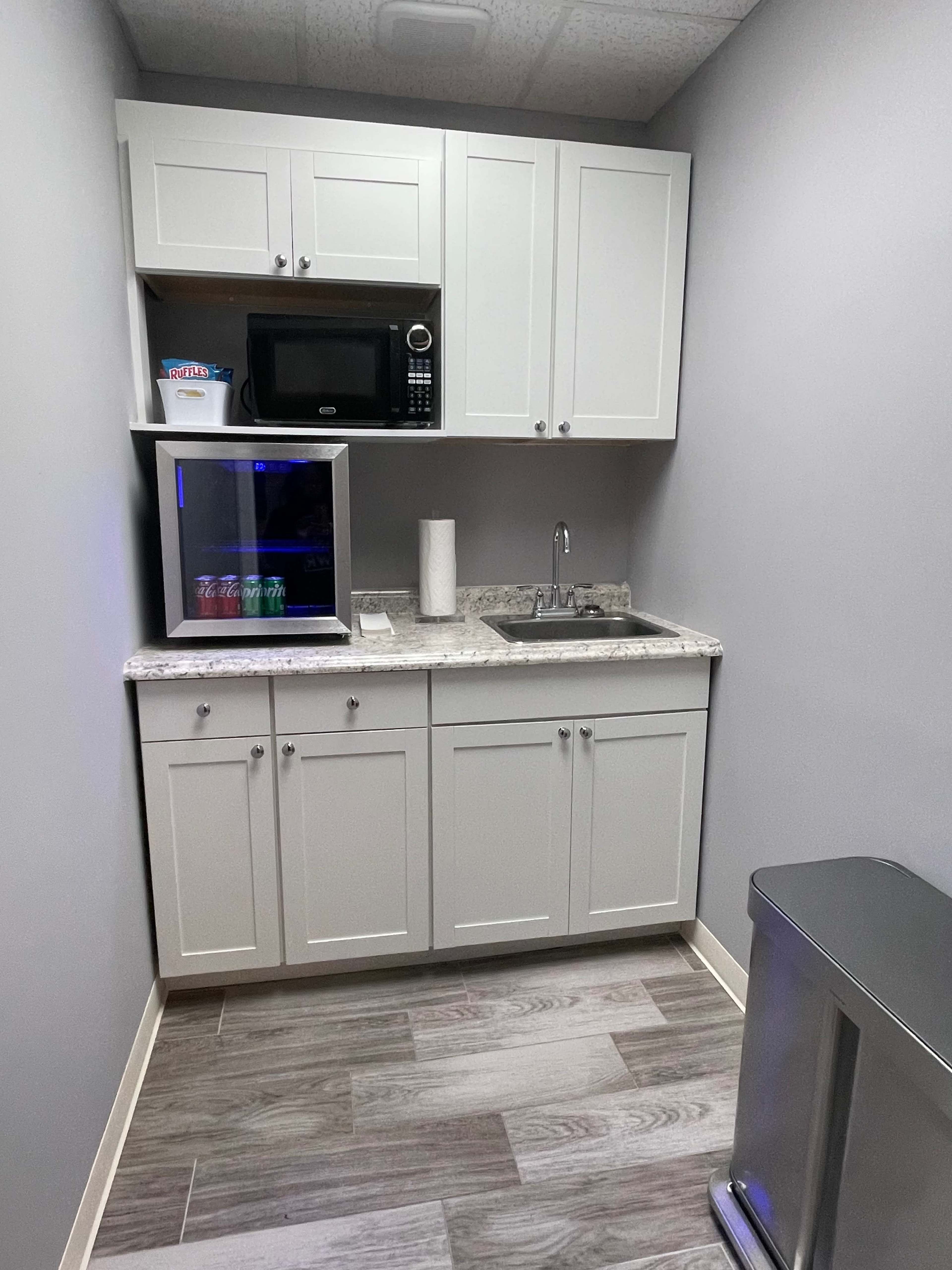 A small kitchenette with a sink, microwave, and a mini-fridge beneath cabinetry, along with a trash can.