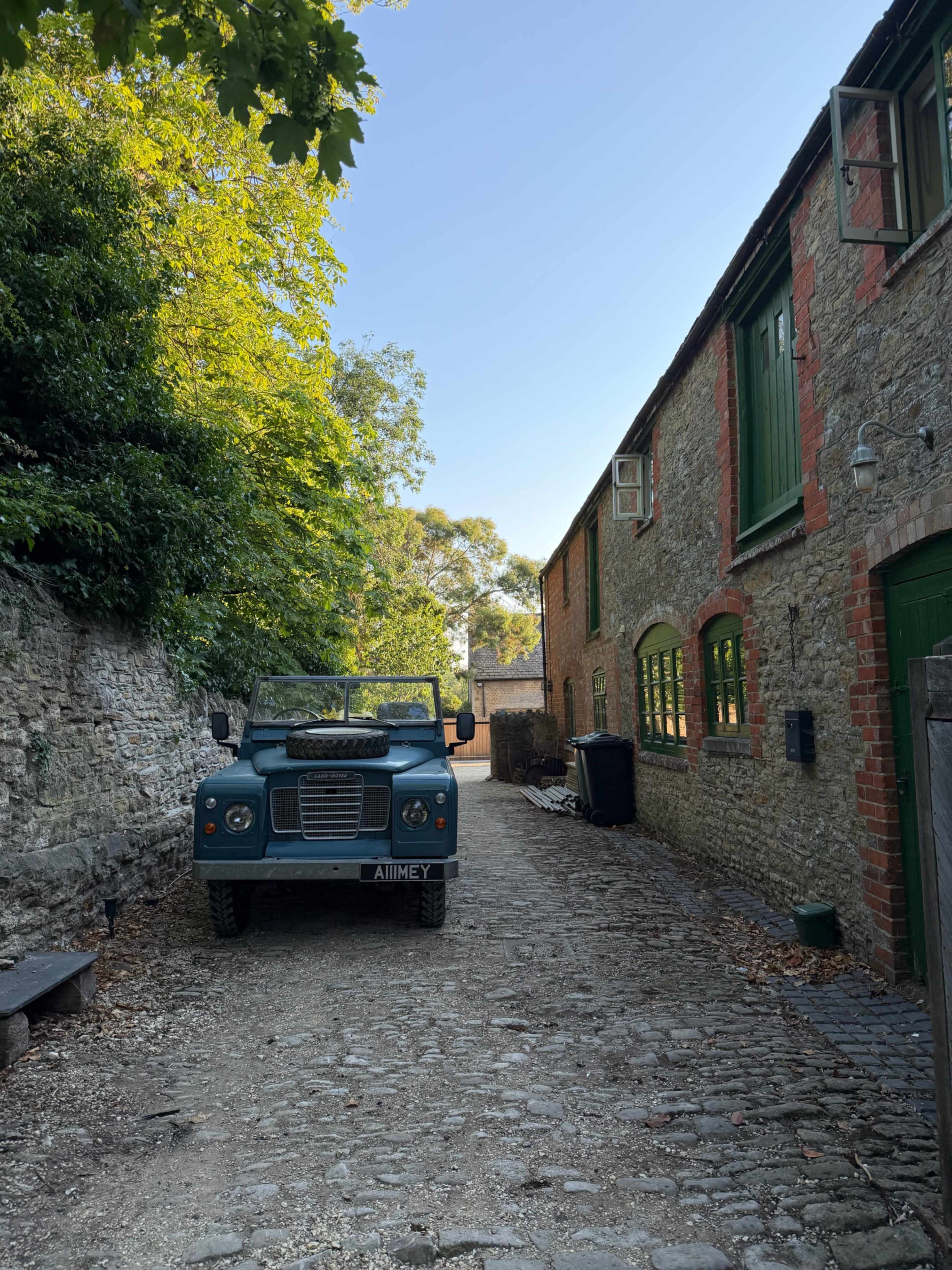 A green Land Rover is parked on a cobblestone path next to brick buildings with open windows and lush trees overhead.