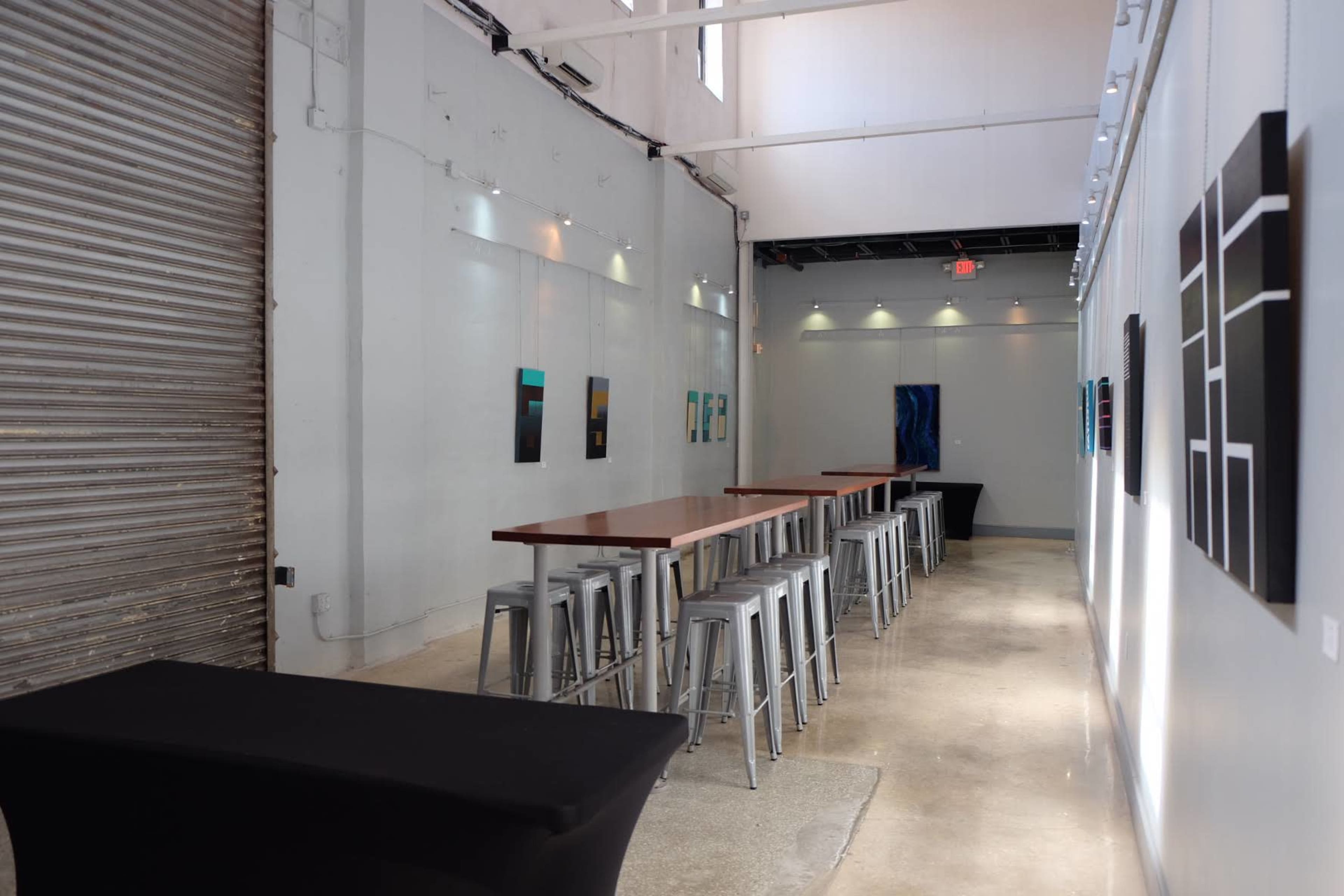 A spacious, modern gallery with metal bar stools and a long table, featuring abstract paintings on the walls.