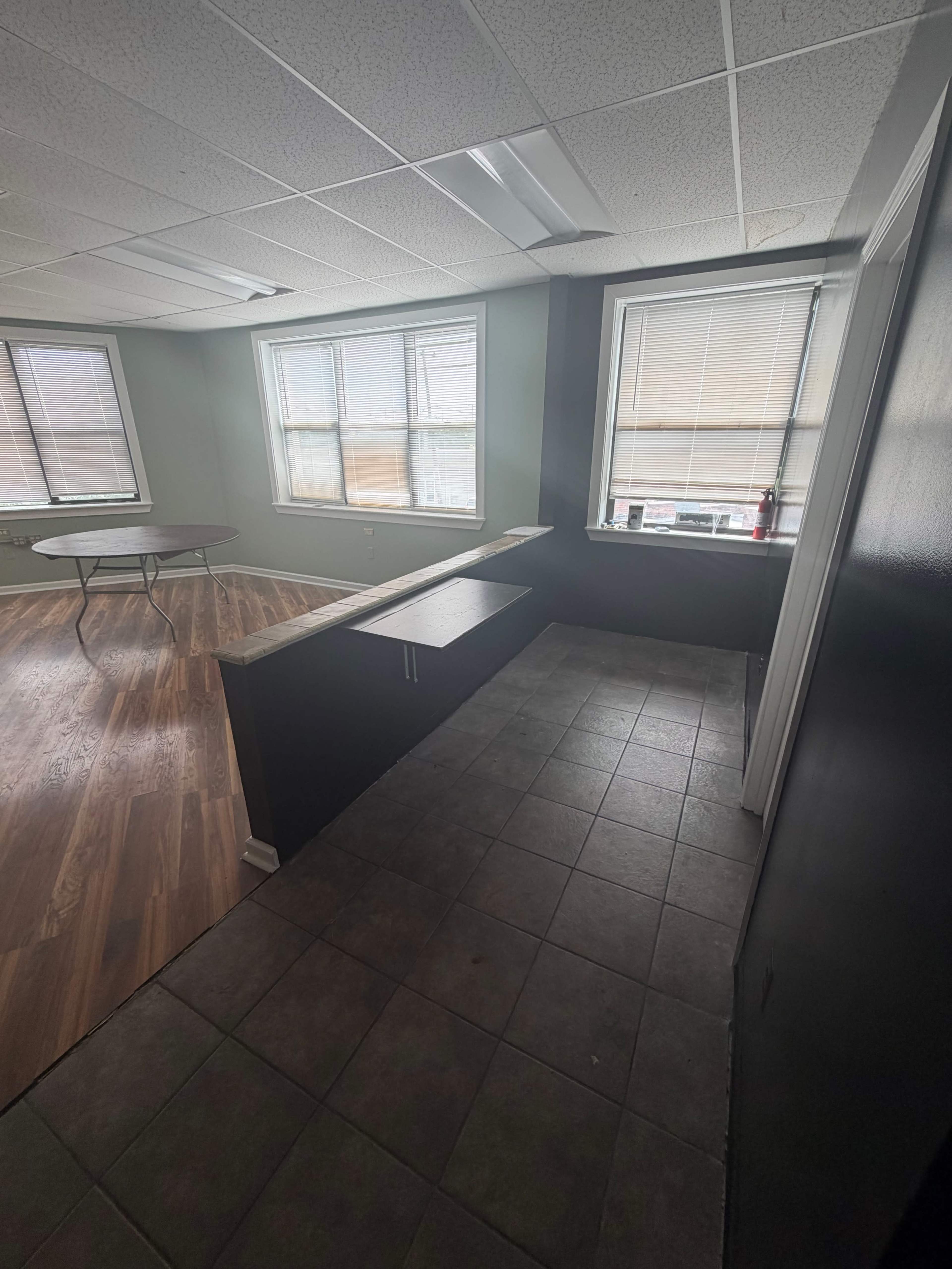 The image shows a partially furnished room with a tiled floor, two large windows, and a countertop along one wall.