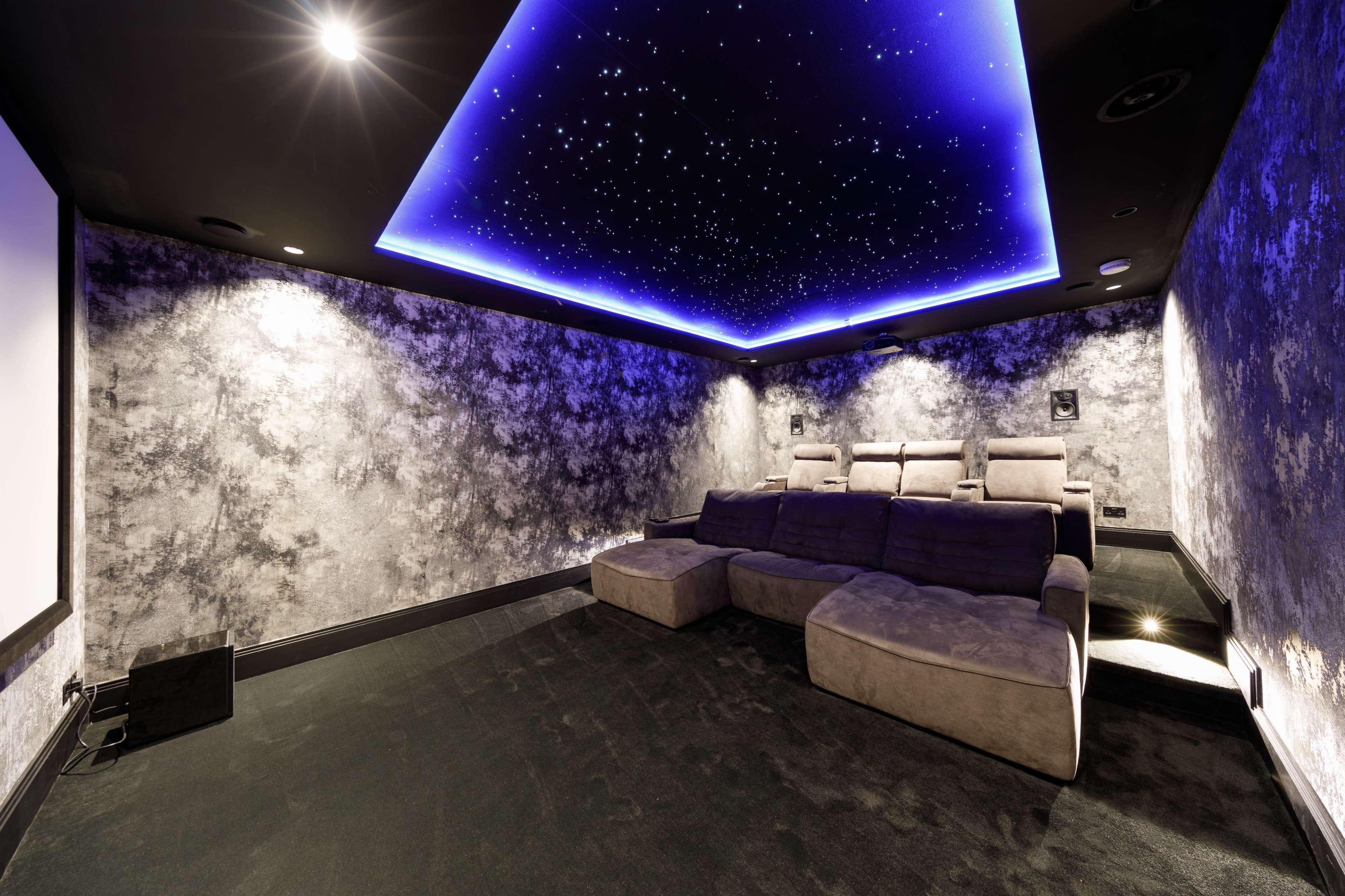 A home theater room with grey textured walls, a starry ceiling, and a large sectional couch arranged for viewing a projection screen.