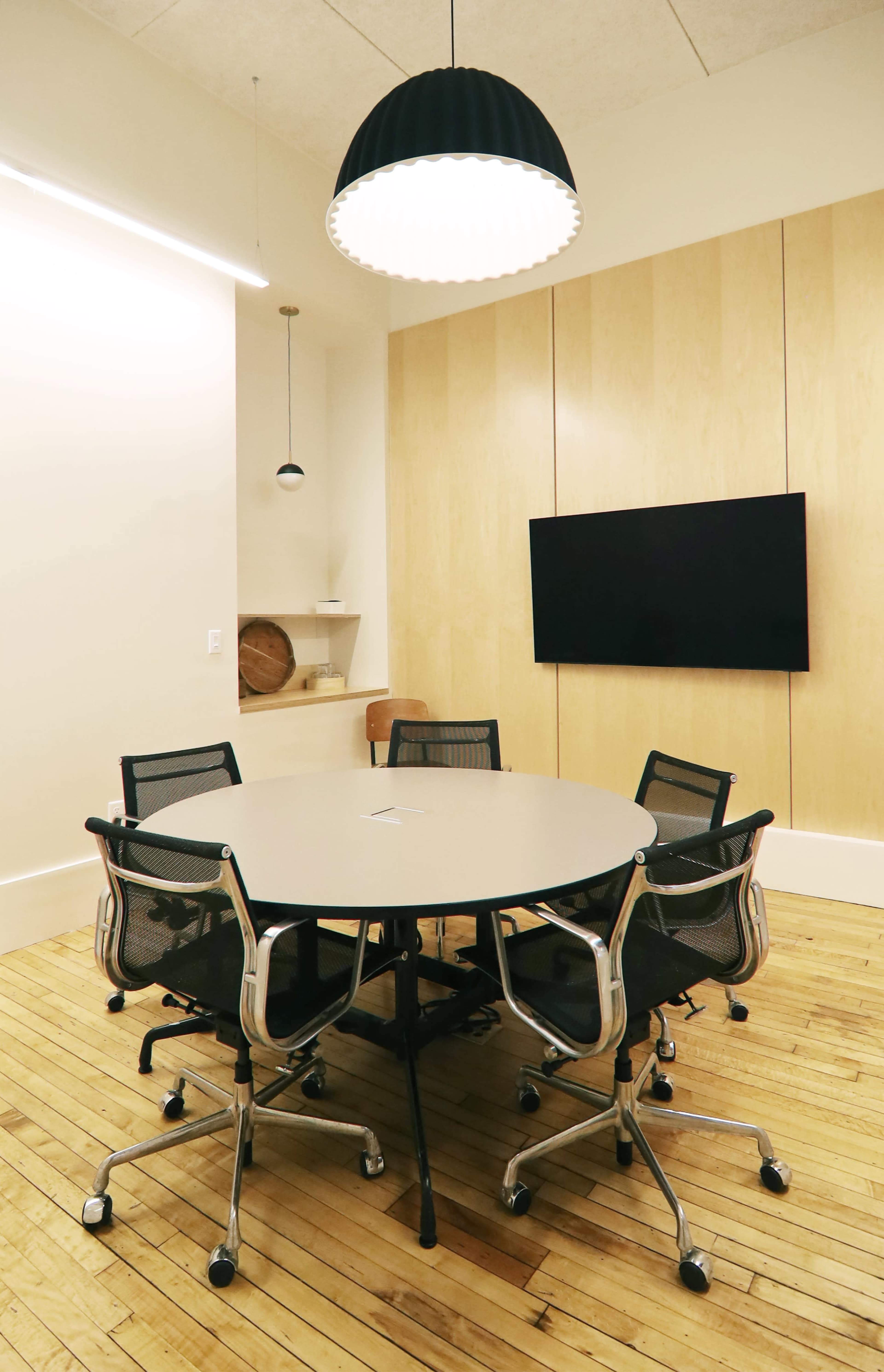 A round conference table surrounded by black mesh chairs is positioned in a minimalist meeting room with wooden flooring and a mounted flat-screen display.
