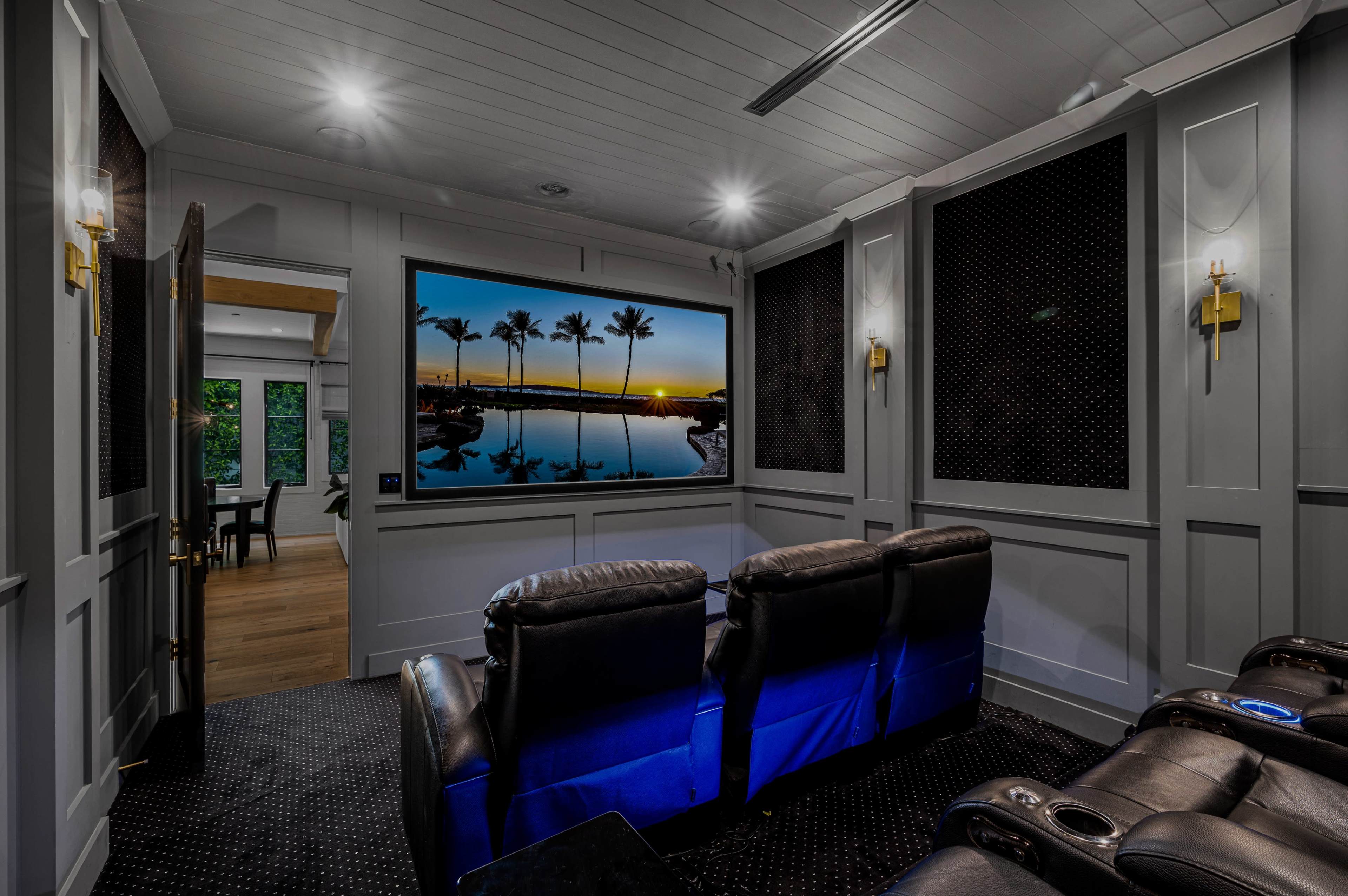 A home theater features recliner seats facing a large screen displaying a sunset view with palm trees reflected in water.