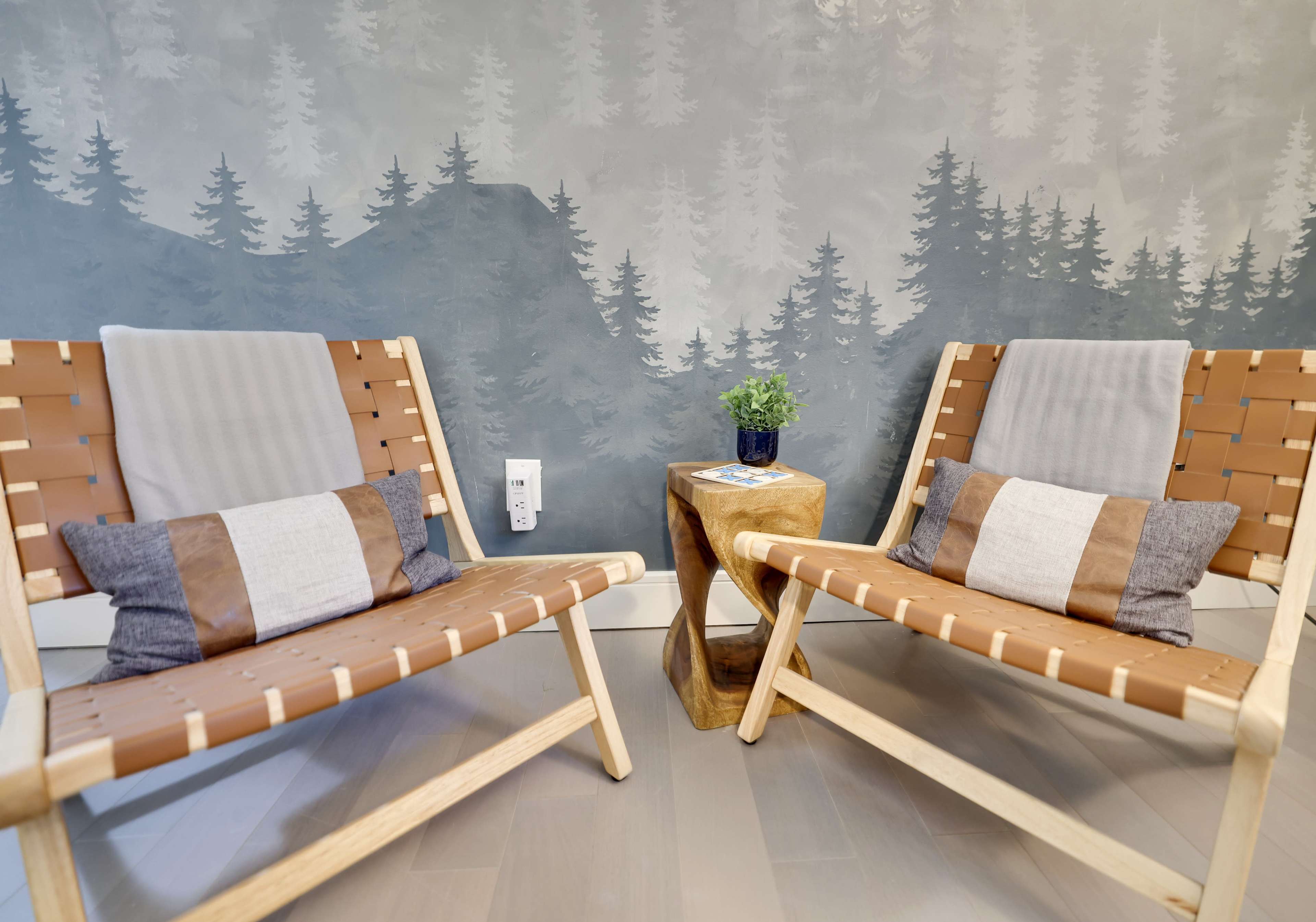 Two modern wooden chairs with woven leather seats are positioned across from each other, flanking a small wooden side table, against a backdrop of a painted mountain and forest mural.