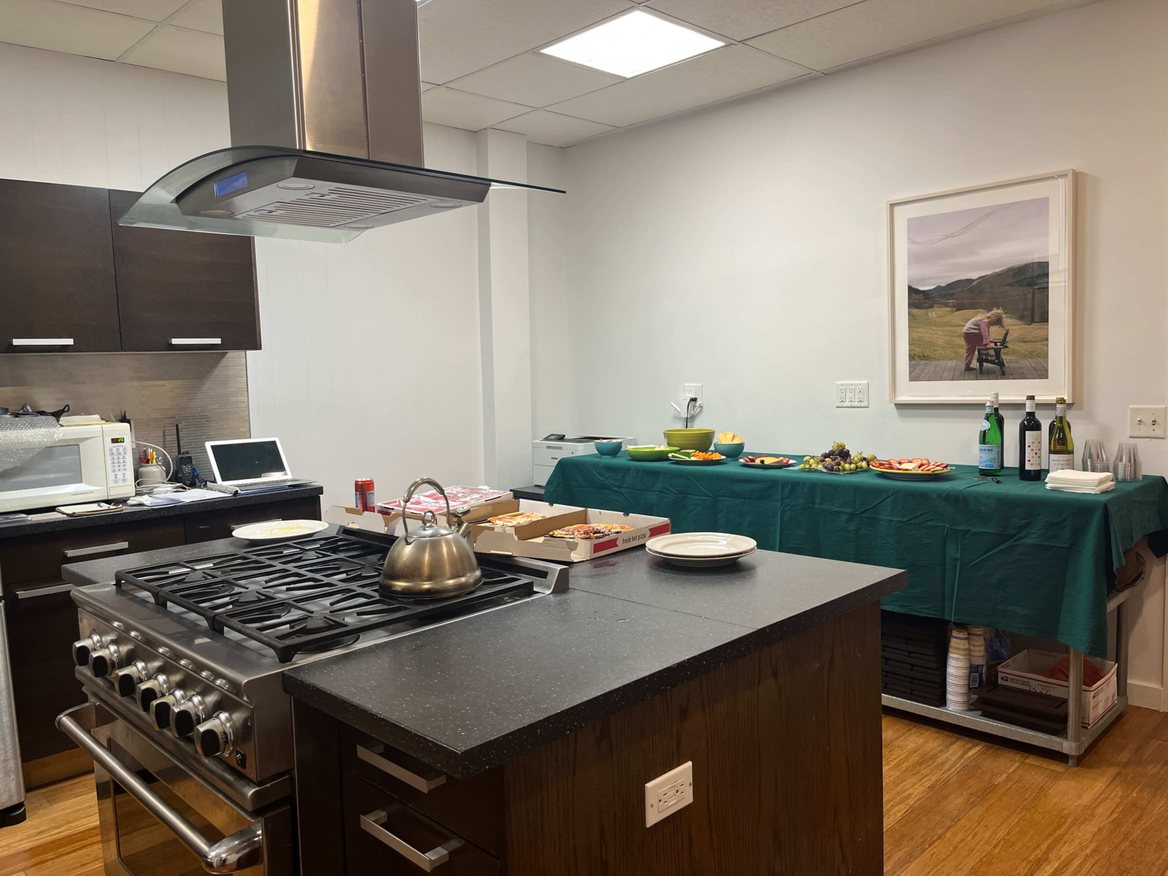 A modern kitchen features a central island with a gas stove, while a table in the background is set with food and drinks against a wall displaying a framed picture.