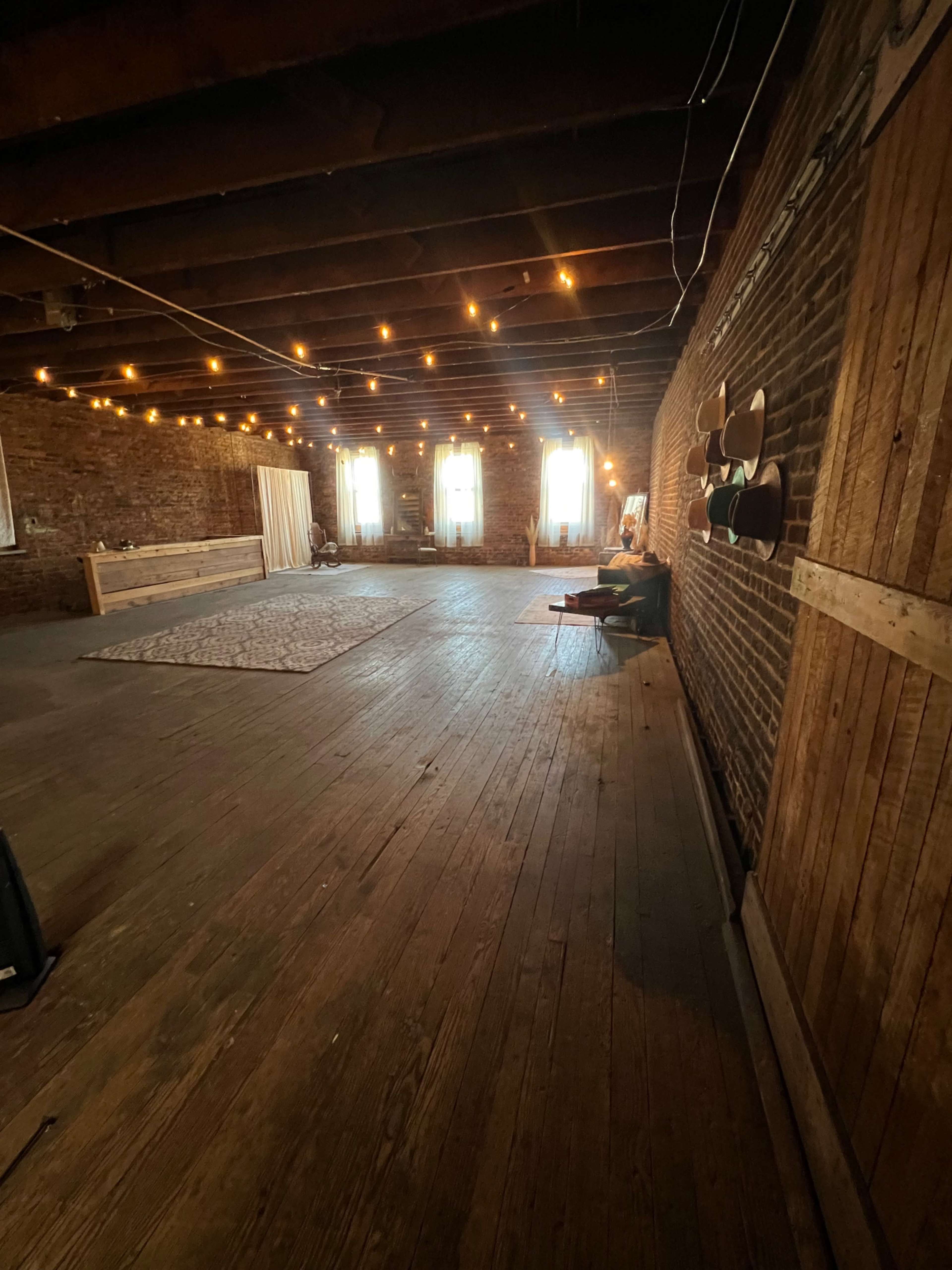The image shows a spacious room with wooden floors, exposed brick walls, and string lights hanging from the ceiling, creating a warm atmosphere.